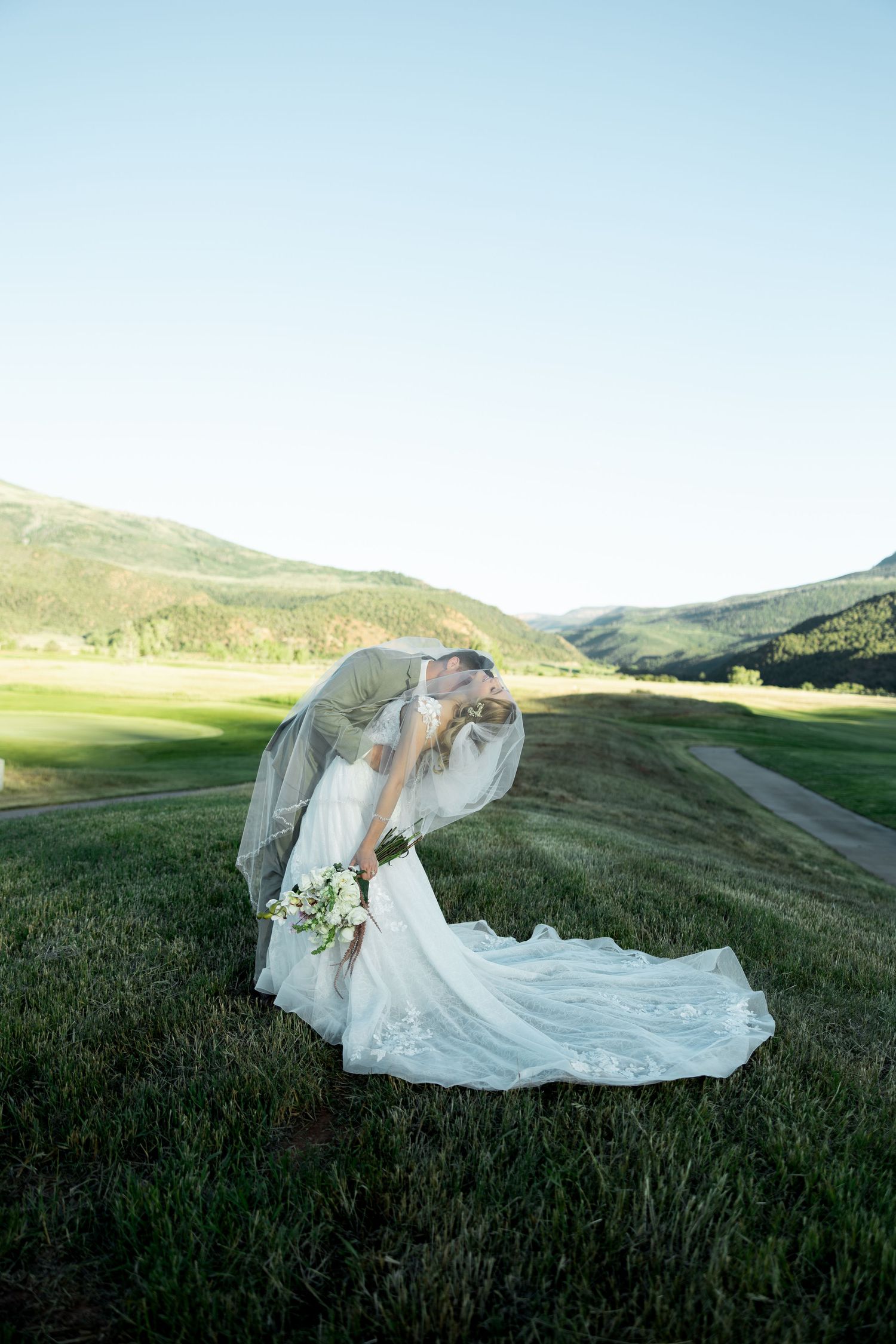 Bride and groom share a romantic dip kiss in a lush green mountain valley, bride in elegant lace gown with long train.