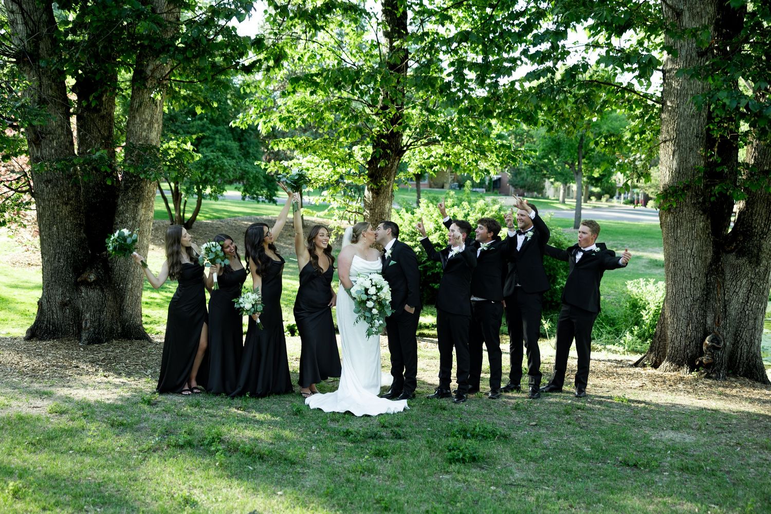 Wedding party in black attire celebrating under lush green trees with bride and groom kissing at center.