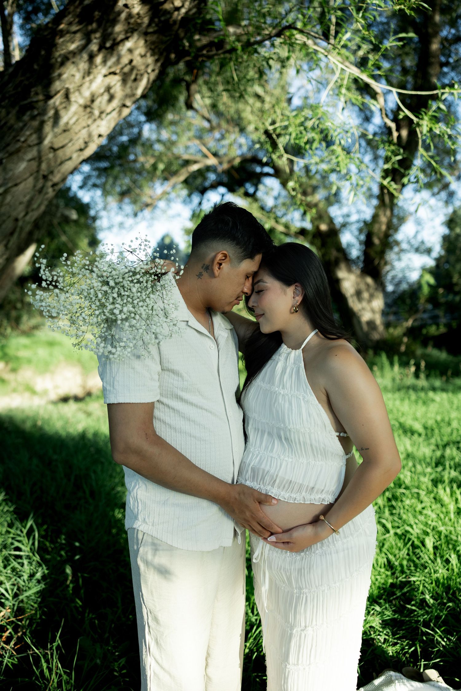 Expecting couple in matching white outfits tenderly touching foreheads outdoors under a large tree in a green park.