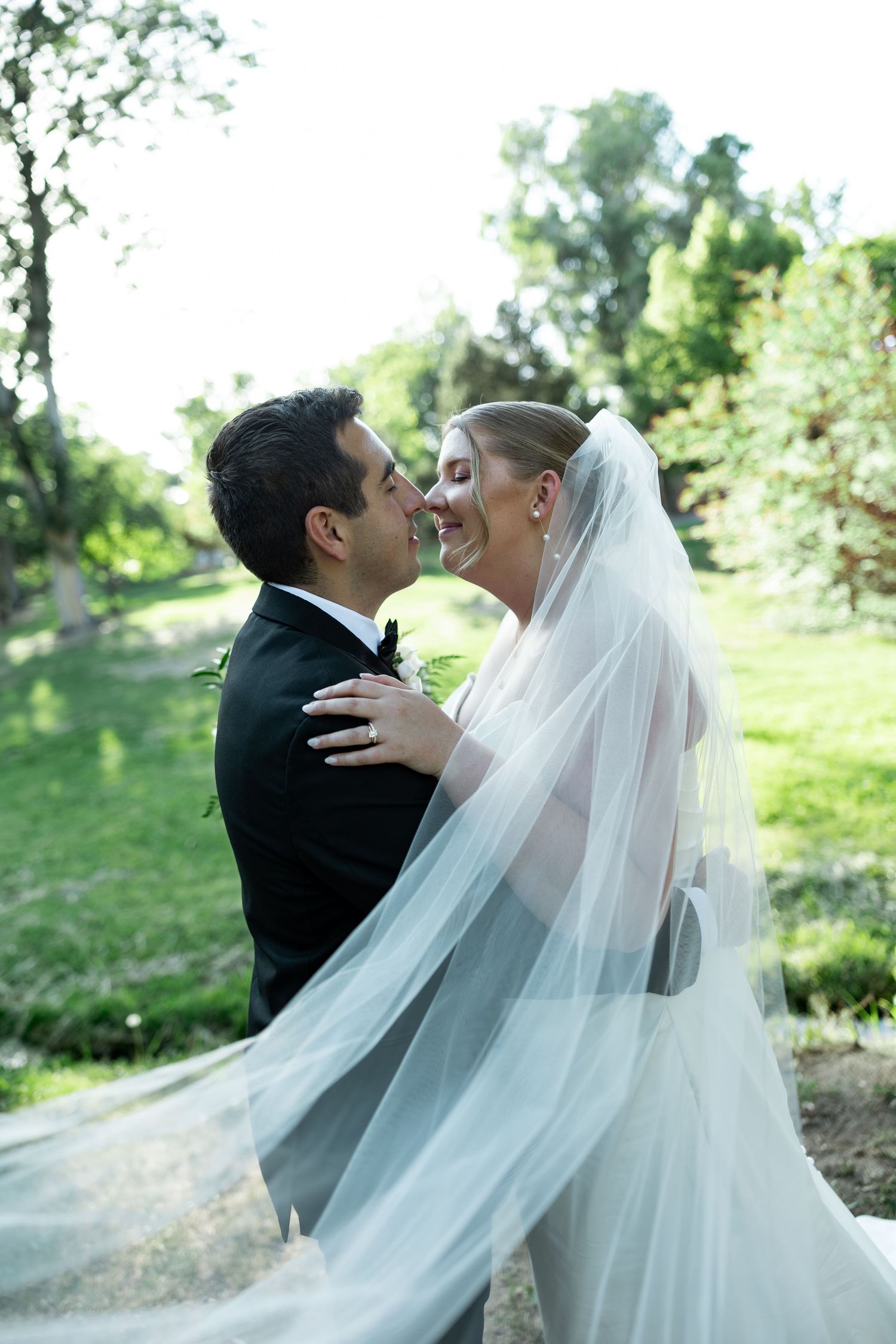 A joyful bride and groom share a romantic moment outdoors, her veil flowing gracefully in a lush green garden setting.