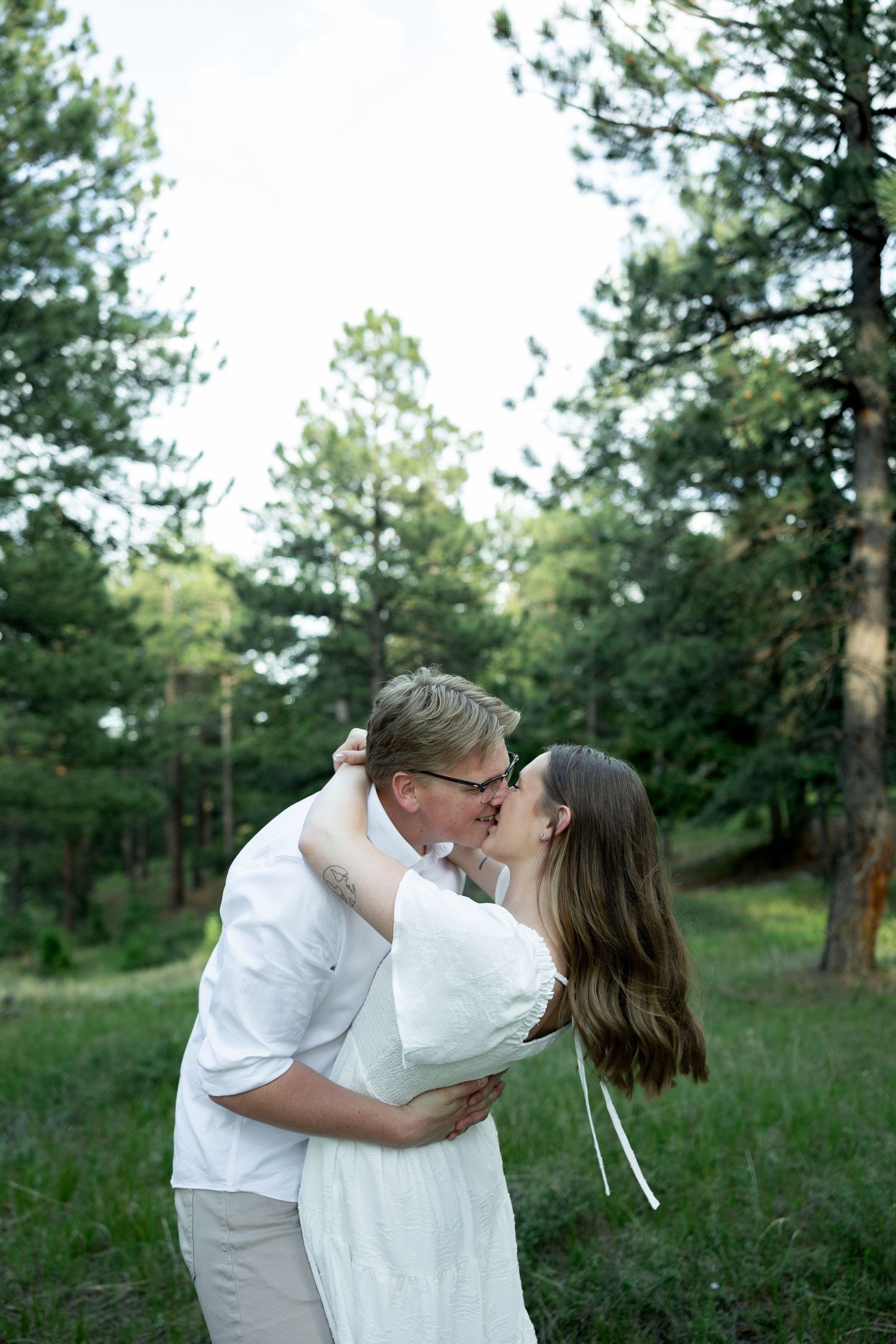 Couple sharing a romantic kiss in a lush green forest clearing, both dressed in white, surrounded by tall pine trees.
