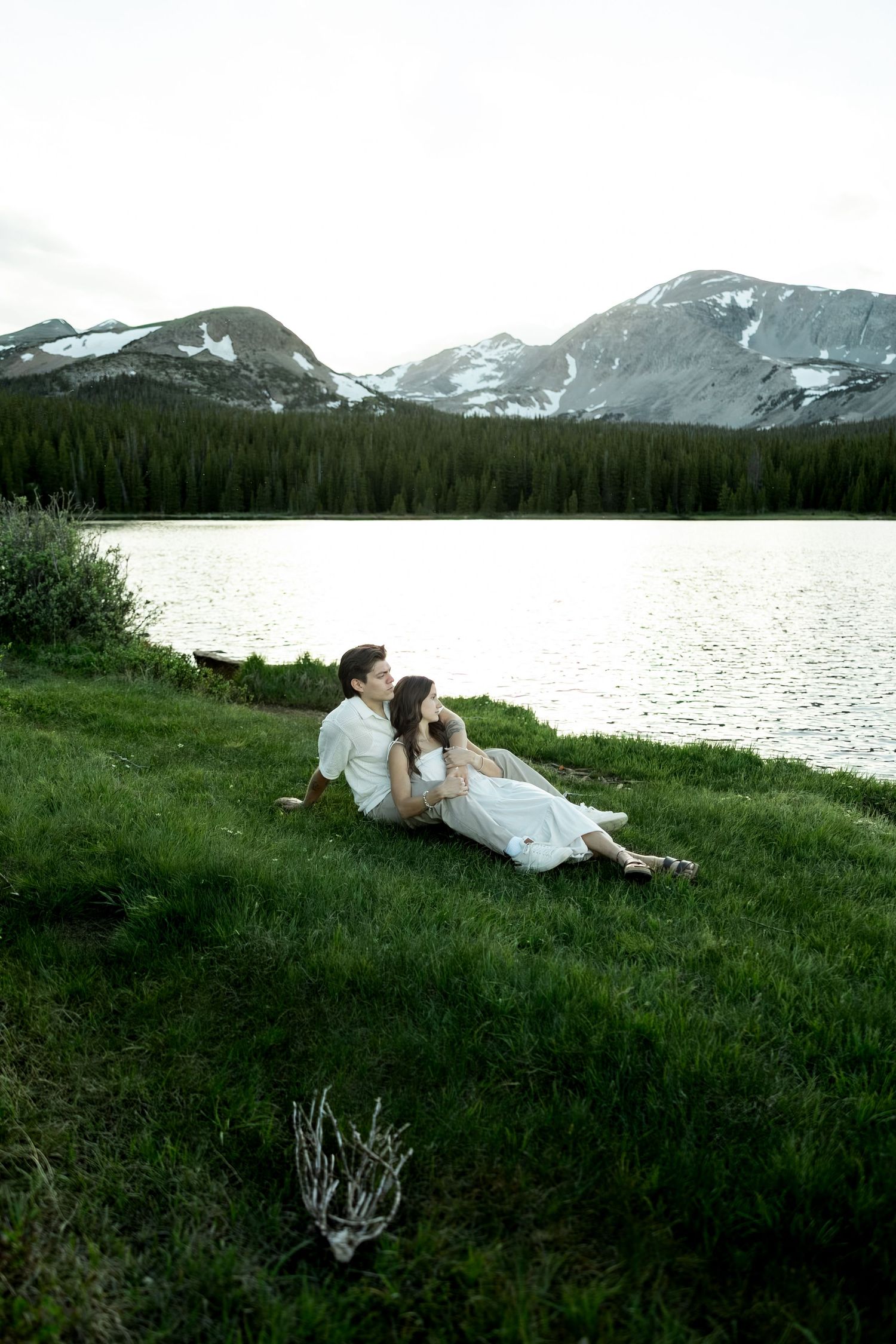Couple sitting together on green grass by a serene mountain lake, surrounded by pine forests and snow-capped peaks.