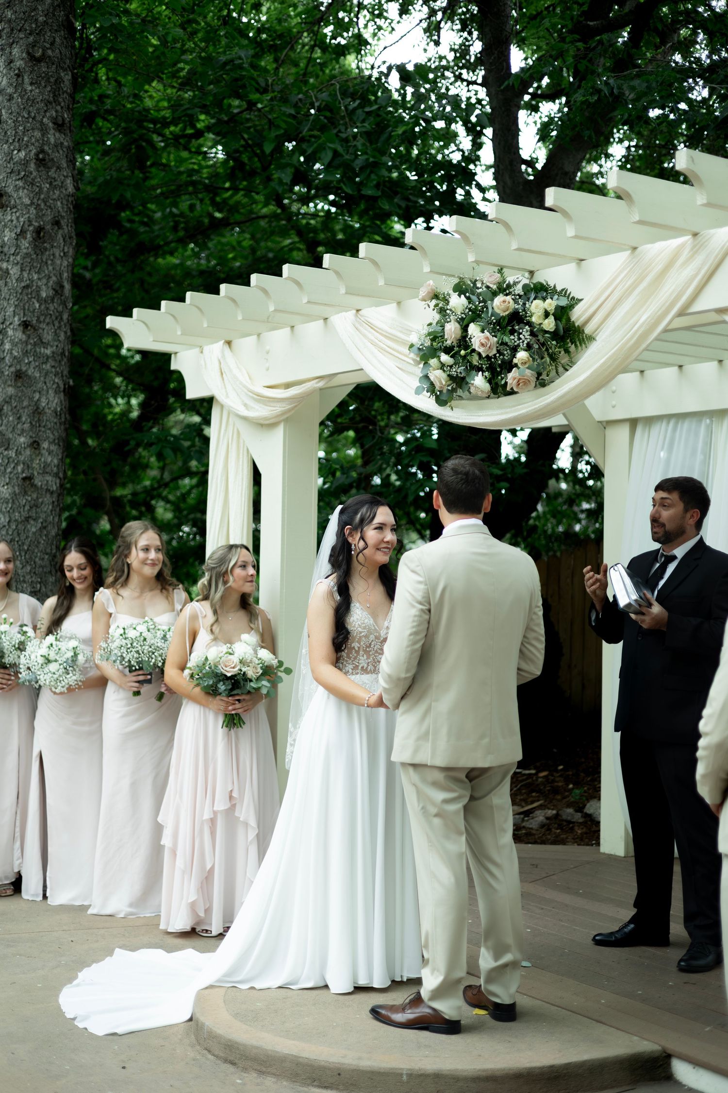 Wedding party lined up under a white pergola decorated with floral arrangements and draped fabric.