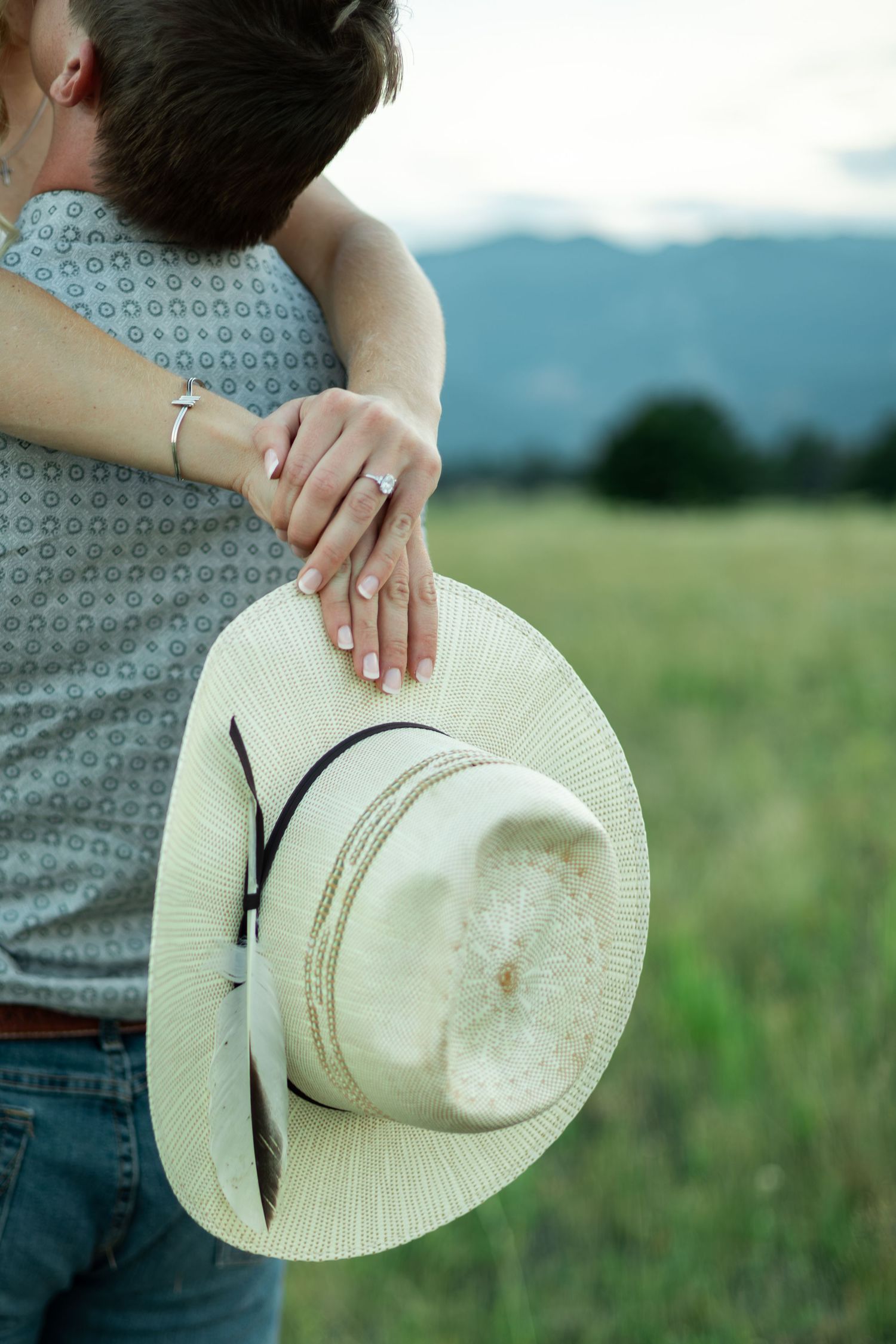 A white cowboy hat is held against a scenic rural background with rolling green hills and mountains.