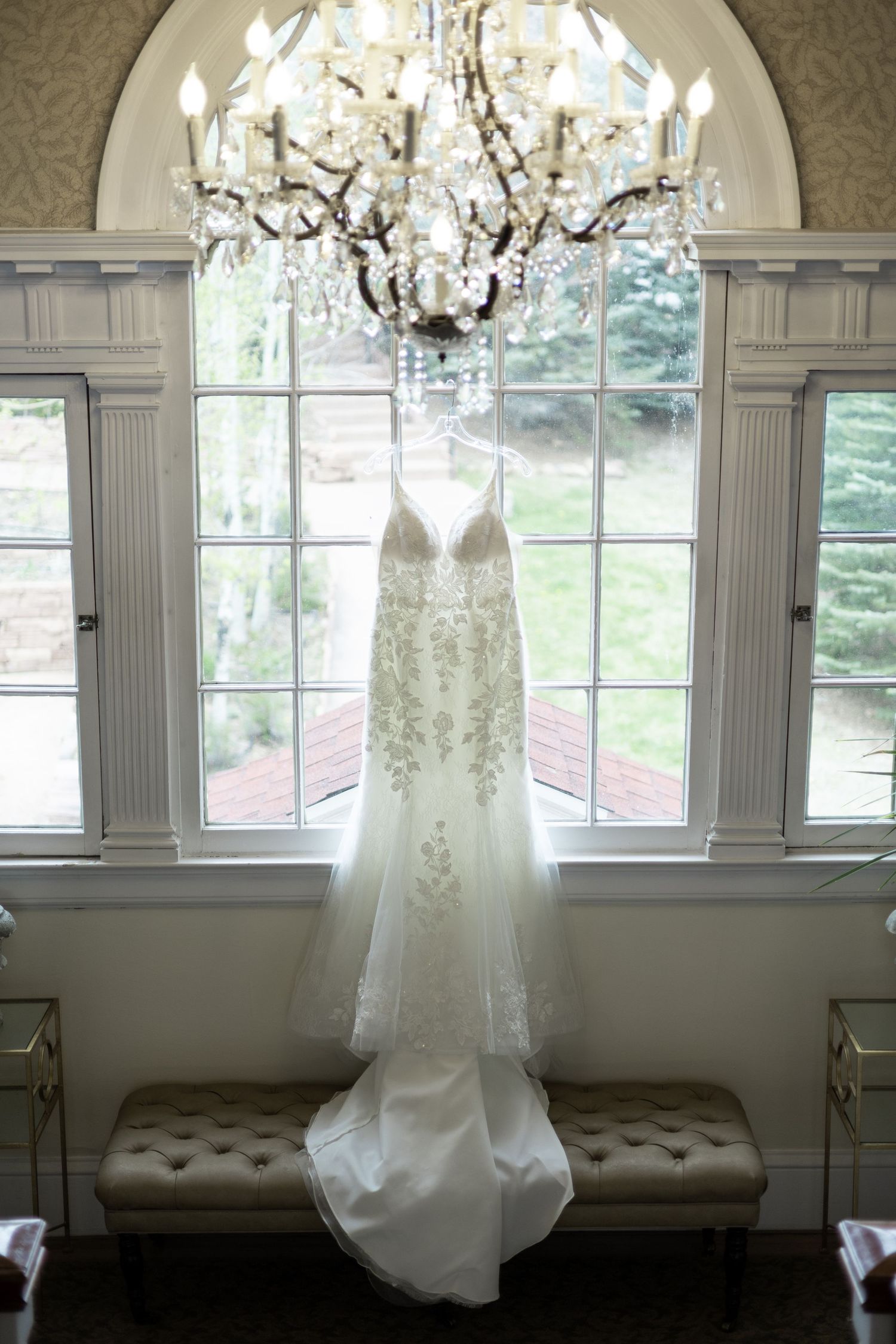 A white lace wedding dress hangs elegantly from a crystal chandelier in front of a large arched window with natural light.