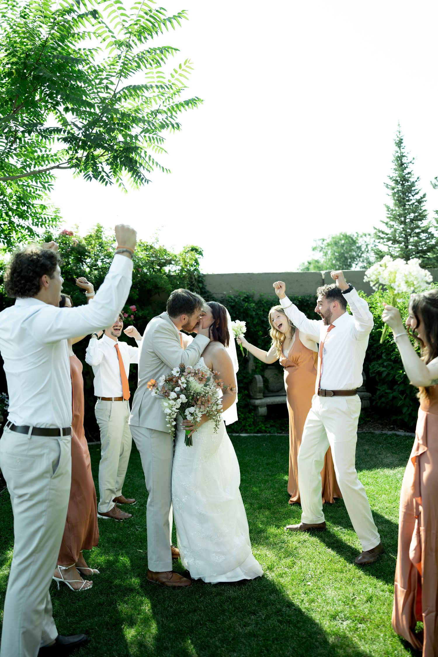 Bride and groom share a kiss surrounded by cheering wedding party on lush green lawn during outdoor ceremony.