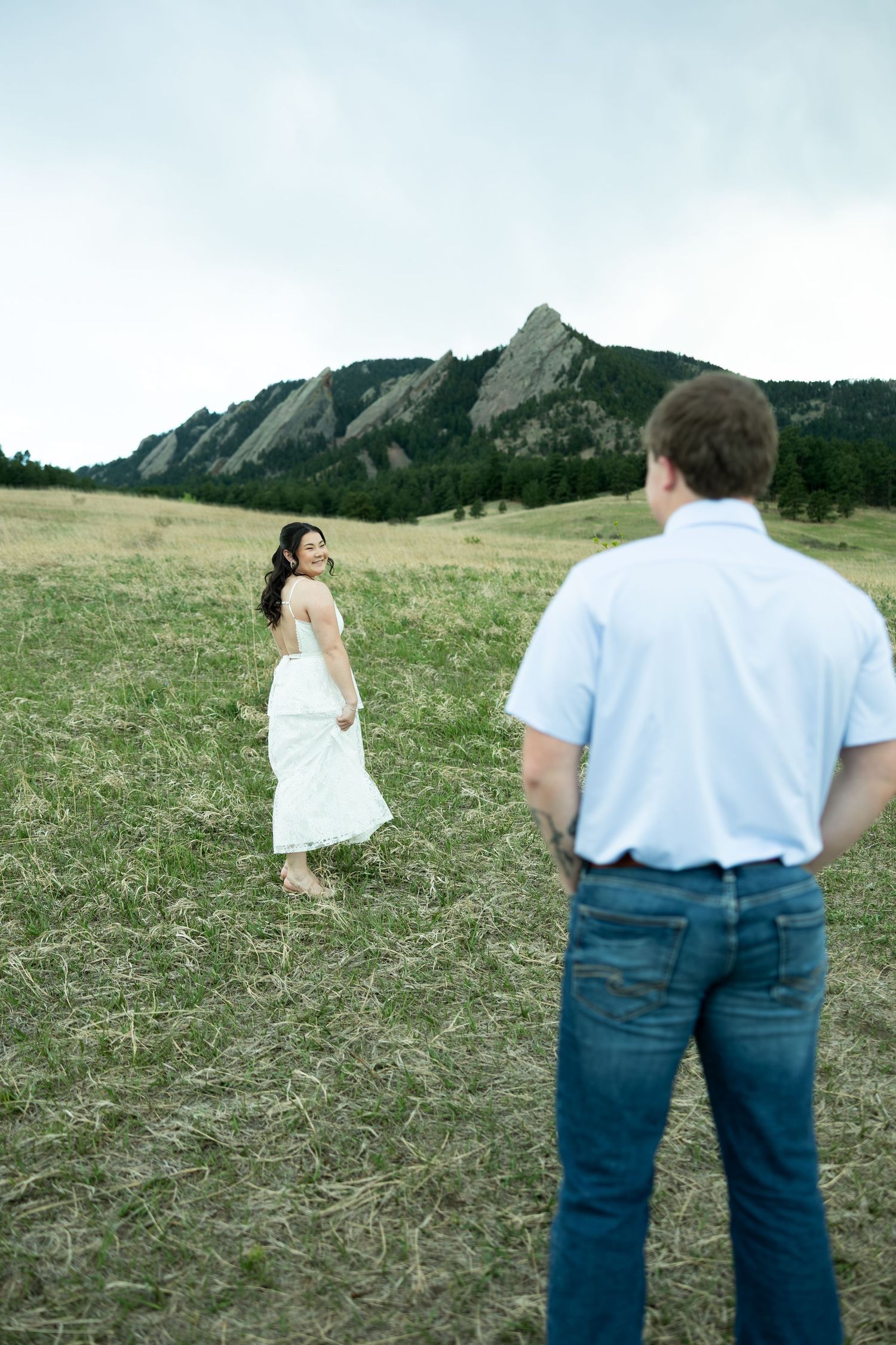 Couple in a grassy field with the Flatirons mountains in Boulder, Colorado, woman in white dress looking back at man.