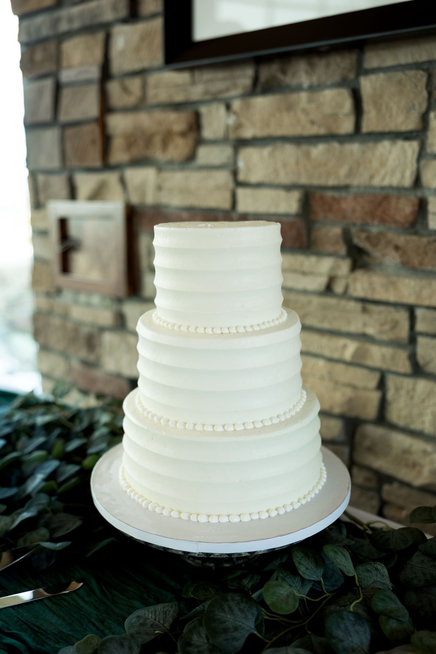 Elegant four-tier white wedding cake with ribbed texture and pearl details displayed against a rustic stone wall.