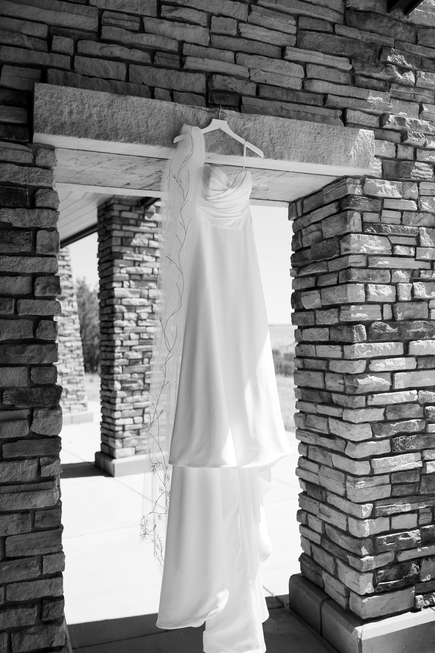Black and white photo of an elegant white wedding dress hanging on a hanger in a rustic stone archway.