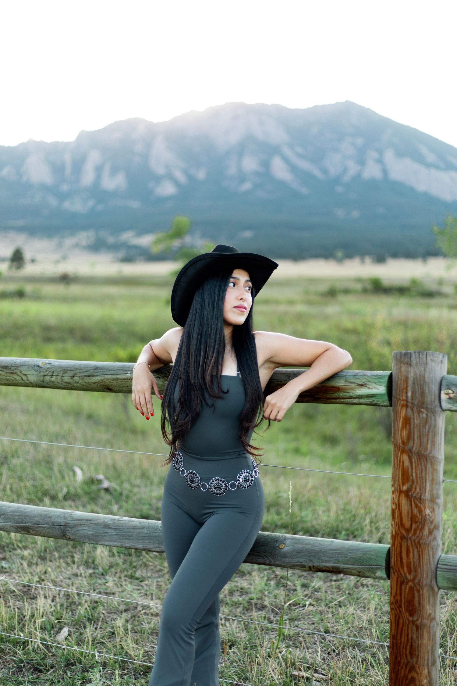 A woman in a black cowboy hat and gray outfit leans on a wooden fence with mountains in the background.