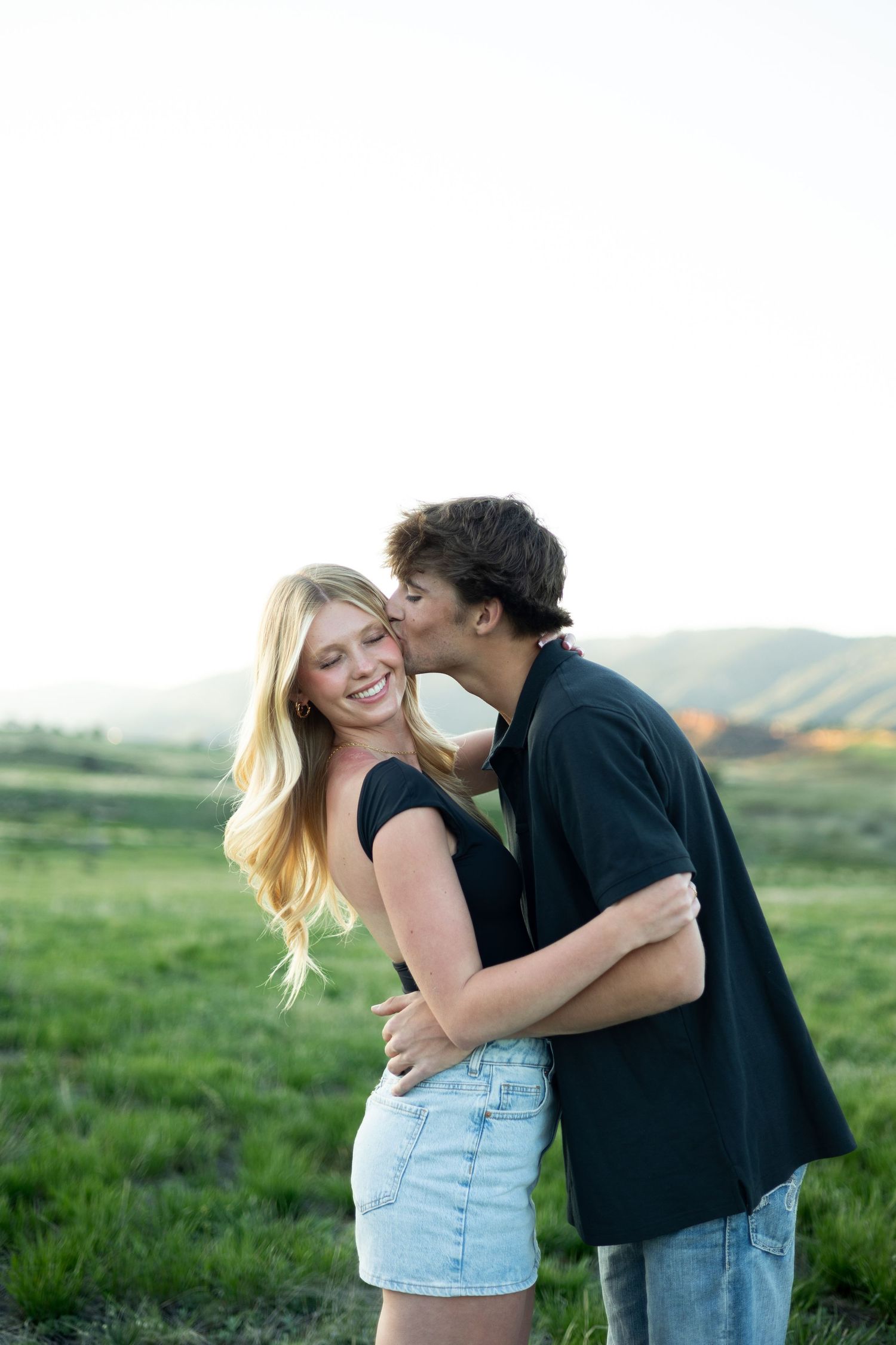 A young couple shares a romantic kiss in a lush green field with rolling hills at golden hour.