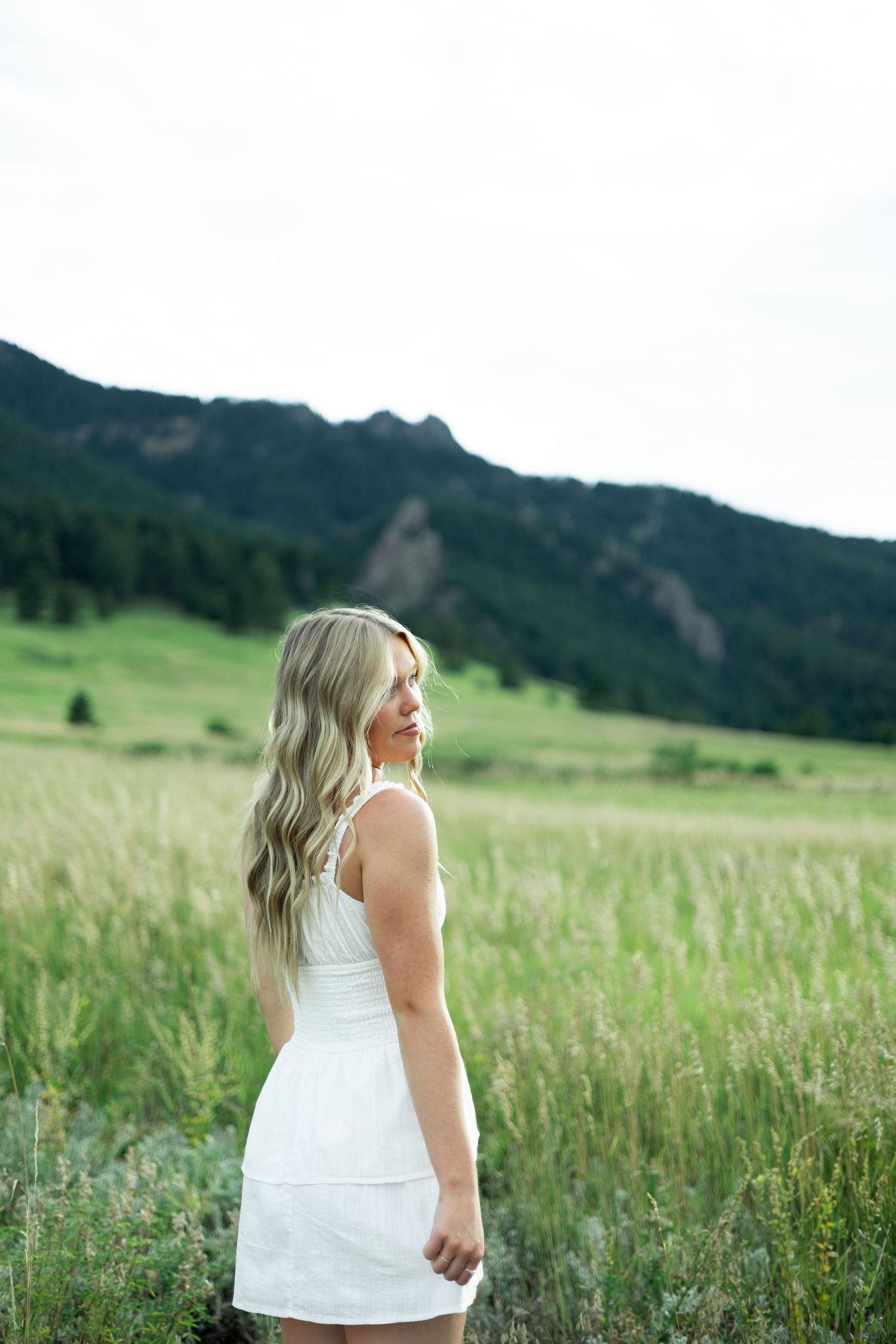 Woman in white dress stands in grassy mountain meadow at dusk.