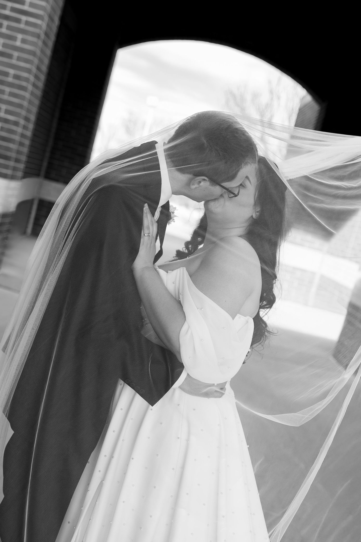 Bride and groom share romantic kiss under flowing veil in black and white photo.