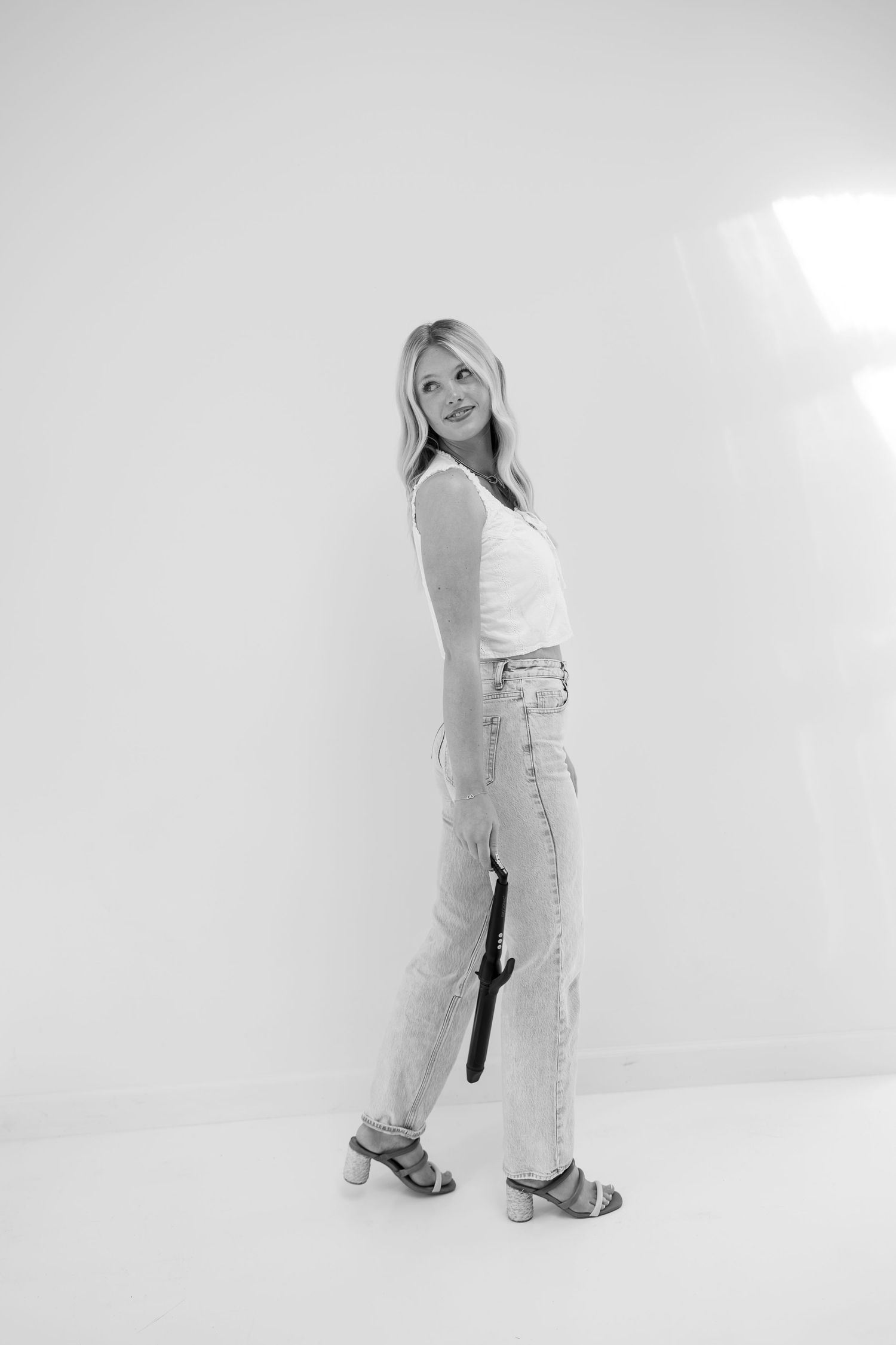 Young blonde woman in white tank top and jeans holding an umbrella, posing in a white studio, black and white photo.