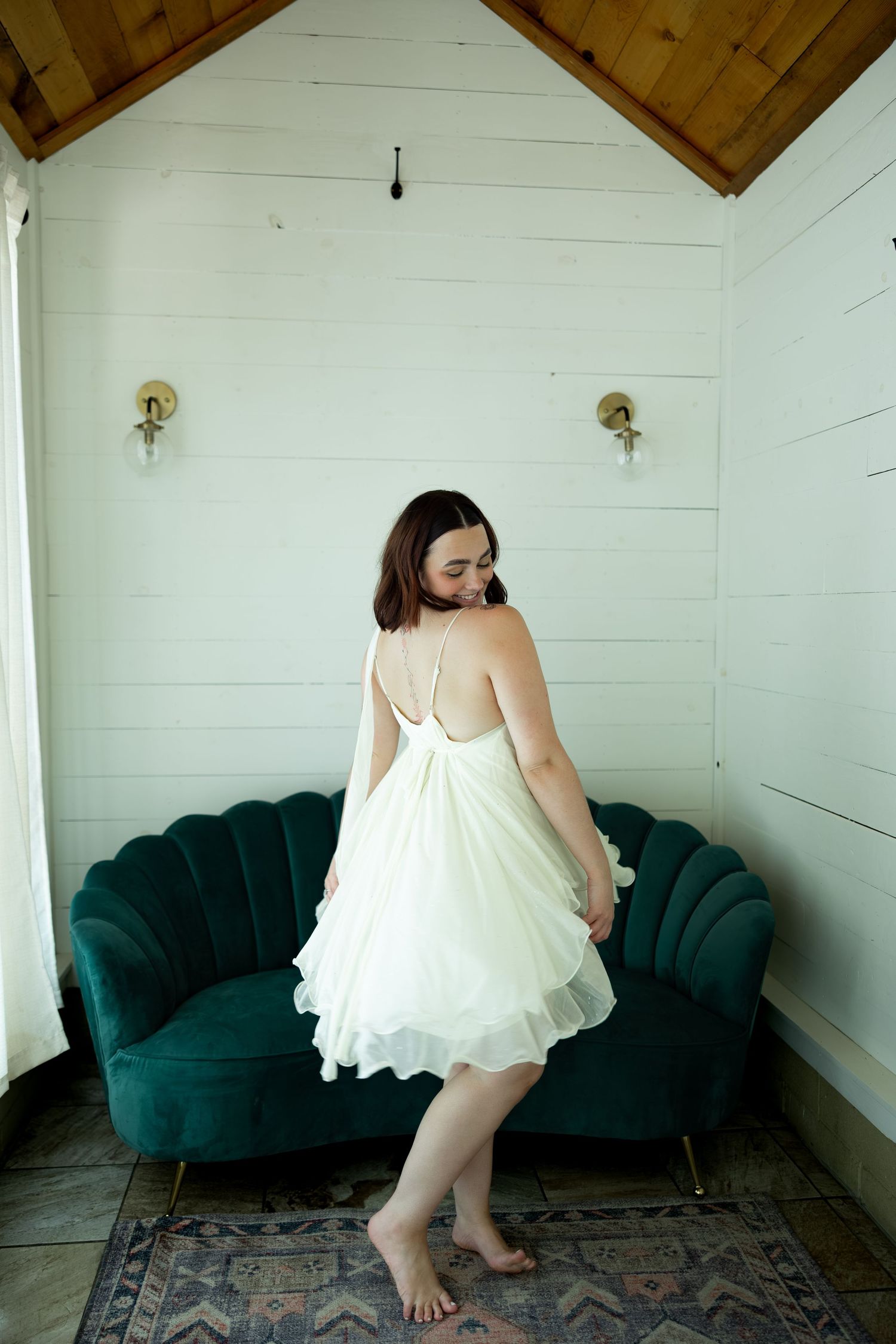 Woman in white chiffon mini dress twirling barefoot in front of green velvet sofa in white shiplap room.