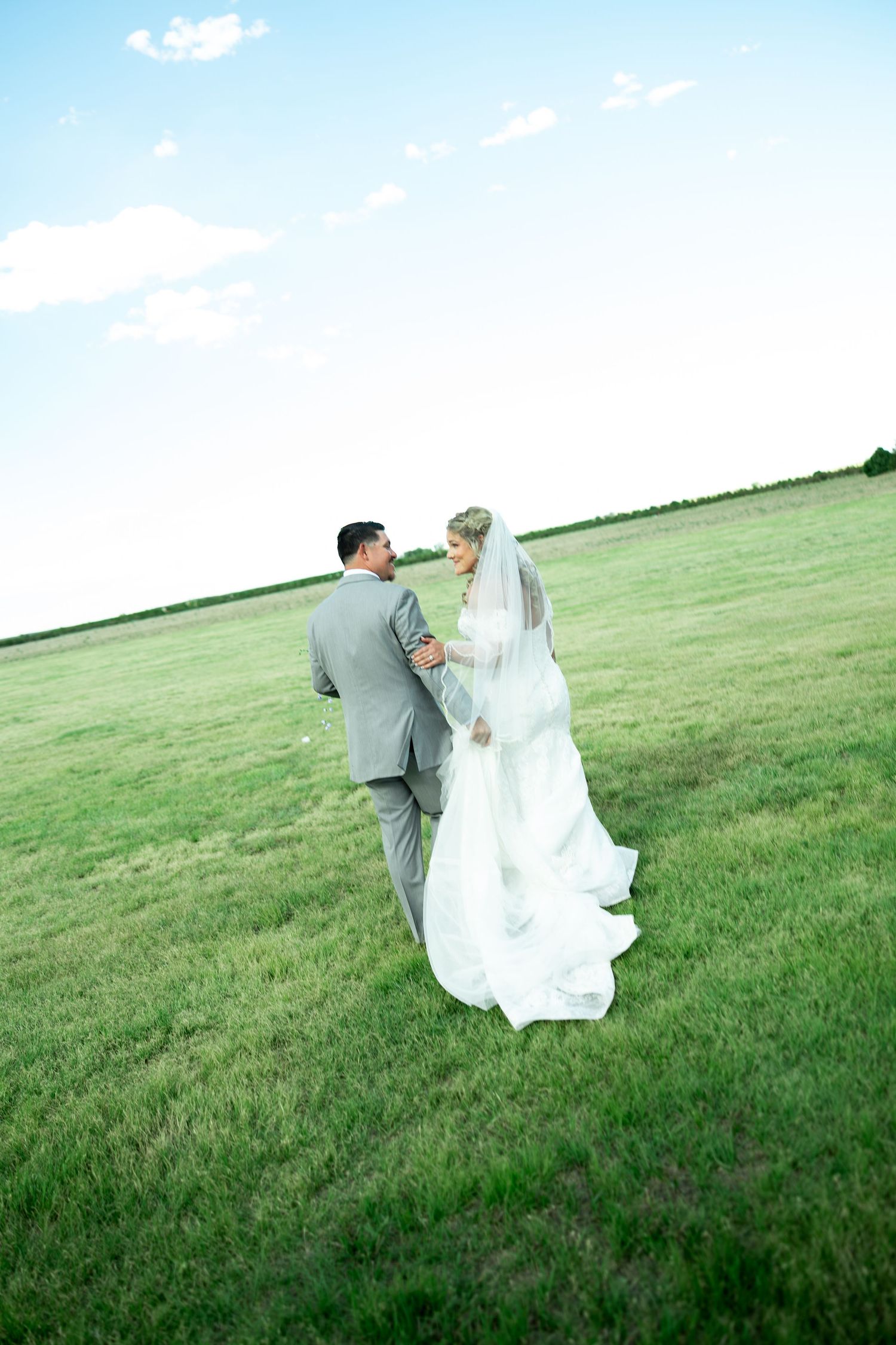 Bride and groom walking hand in hand across a lush green field, sharing a romantic glance on their wedding day.