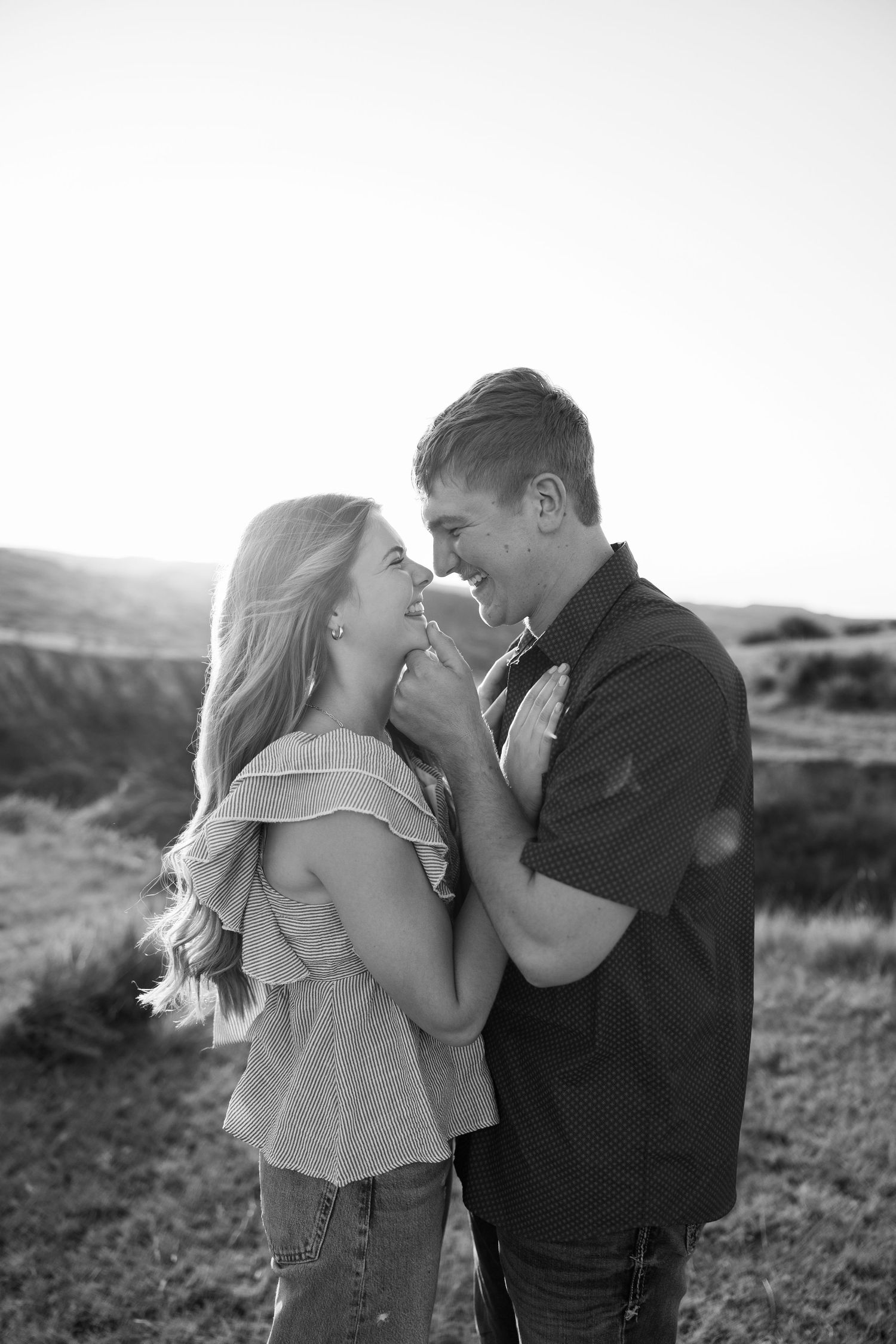 Black and white photo of a smiling couple holding hands and gazing at each other in a scenic outdoor field.