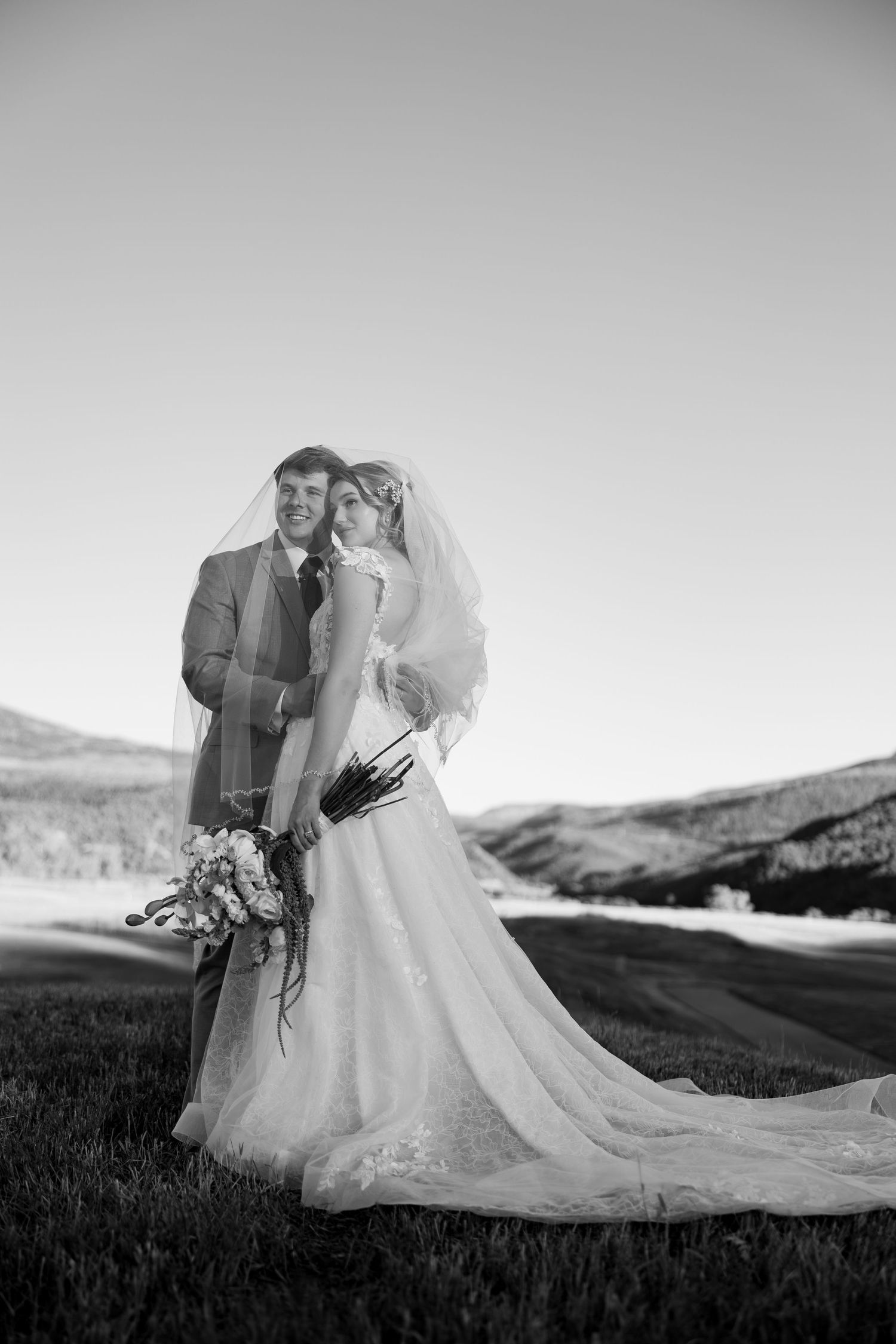 Black and white photo of a bride and groom embracing outdoors with mountain landscape backdrop on their wedding day.