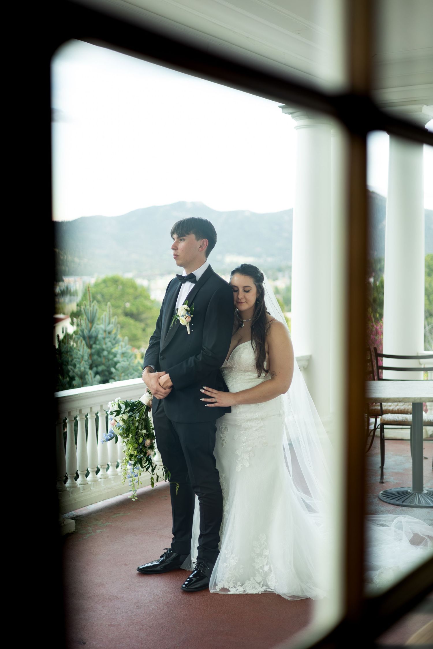 A couple on their wedding day stands on a covered porch with mountain views, viewed through a window frame.