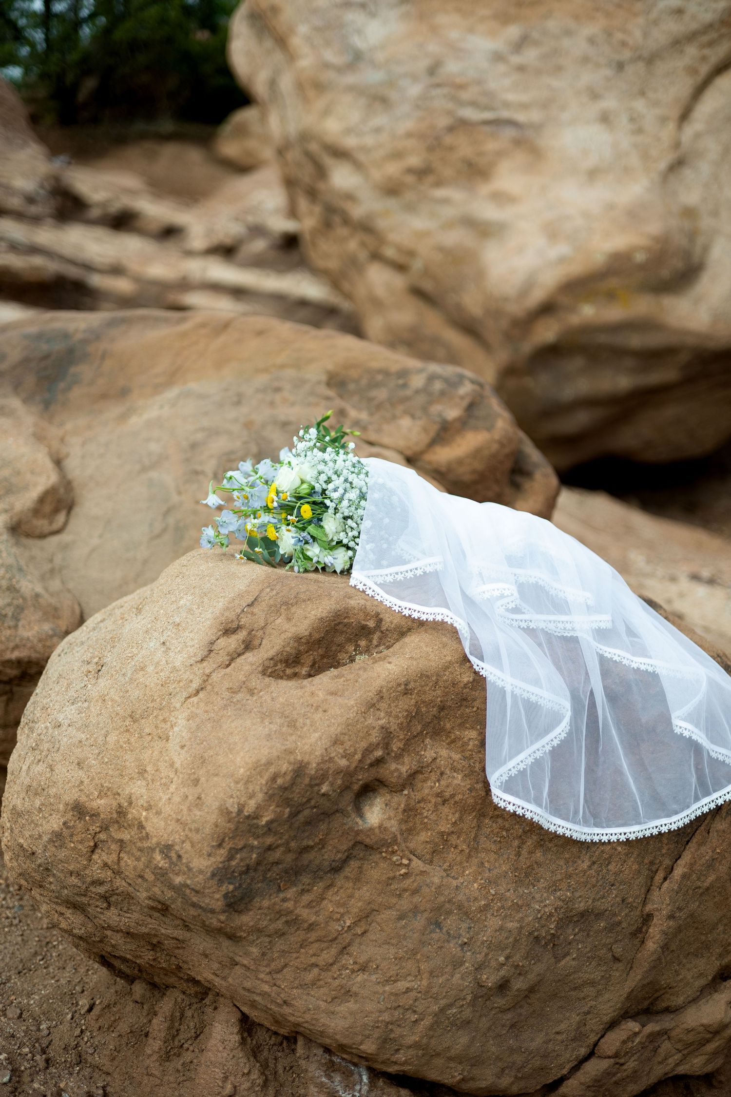 A white bridal veil with delicate floral accents blows gently in the wind while draped over natural brown rocks outdoors.