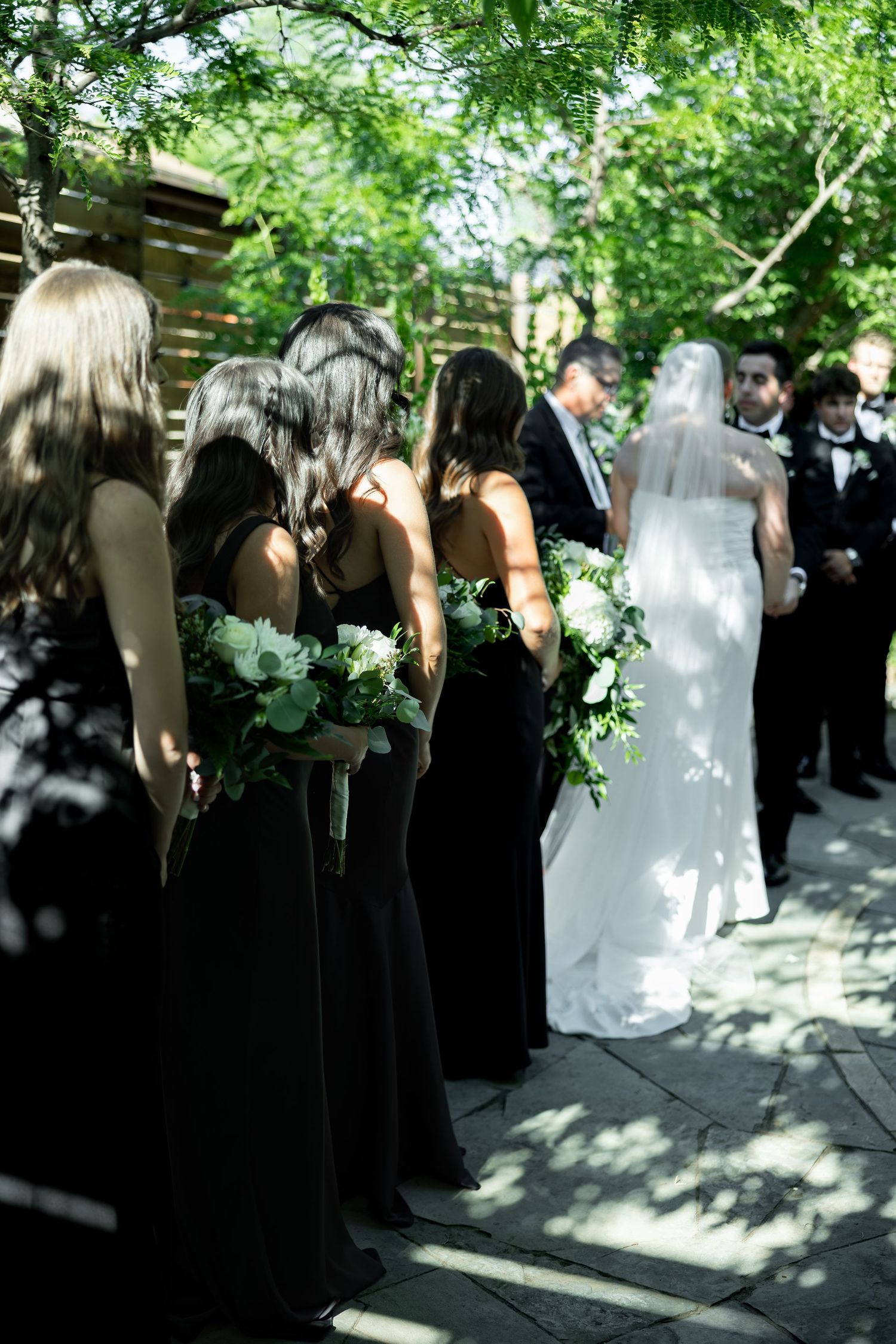 Bridesmaids in black gowns holding white and greenery bouquets stand beside a bride in white veil at an outdoor wedding ceremony.