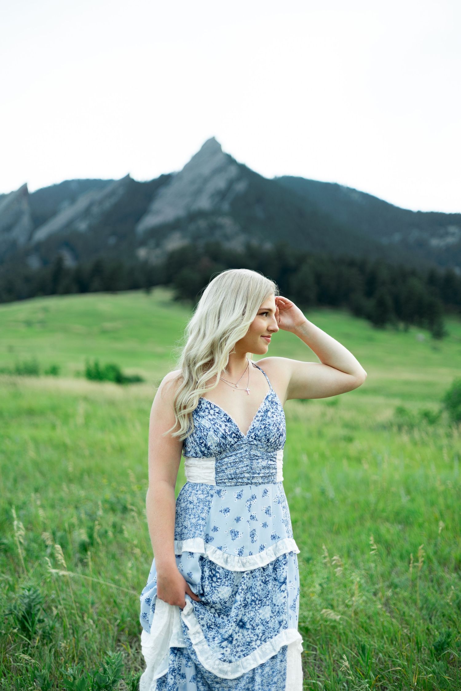 Woman in blue floral dress standing in lush green meadow with dramatic mountain landscape backdrop.