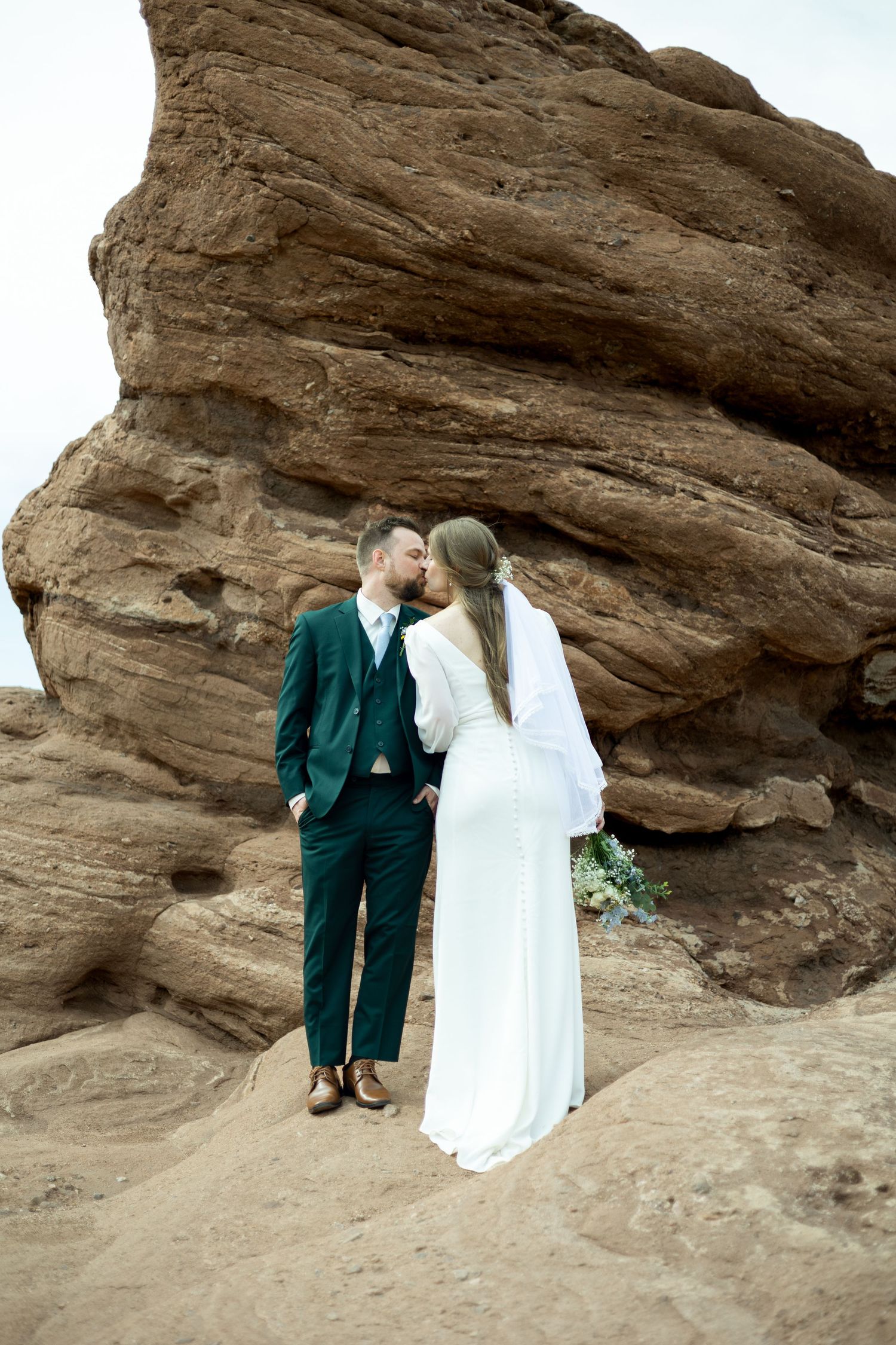 Bride and groom kissing at Red Rocks, Colorado, wearing white gown and green suit against red sandstone formations.