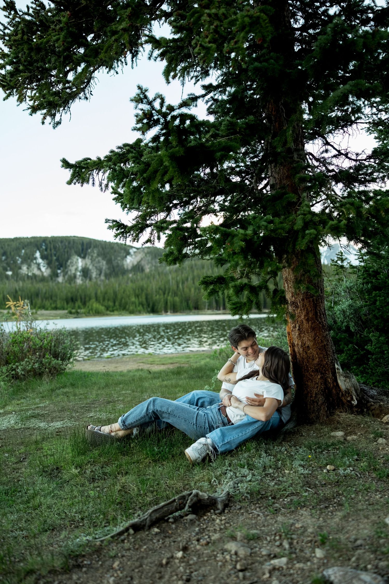Couple sitting under a pine tree by a serene mountain lake, embracing in a lush green landscape.