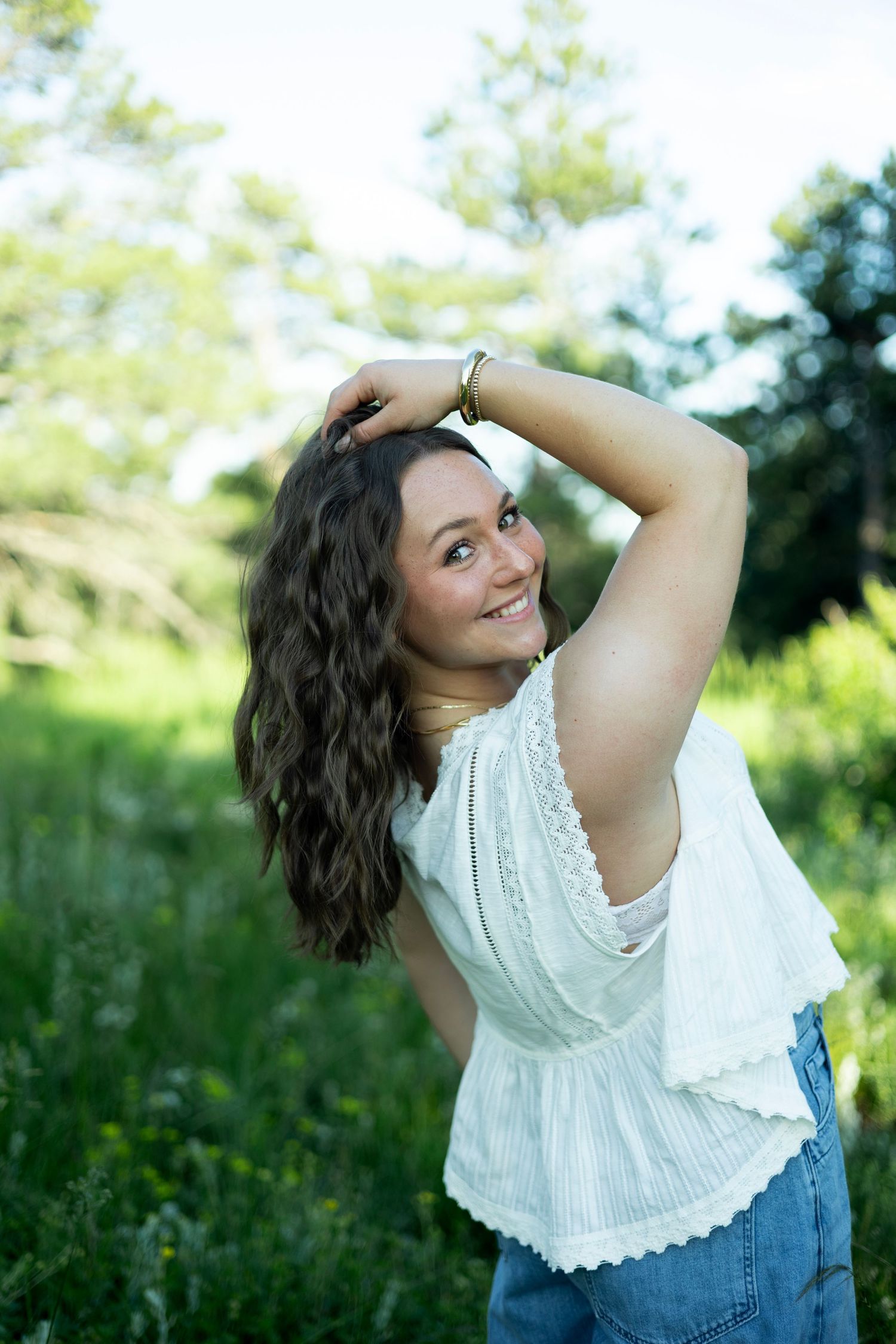 Young woman with curly brown hair smiling outdoors in a white top and jeans, posing in a lush green field.