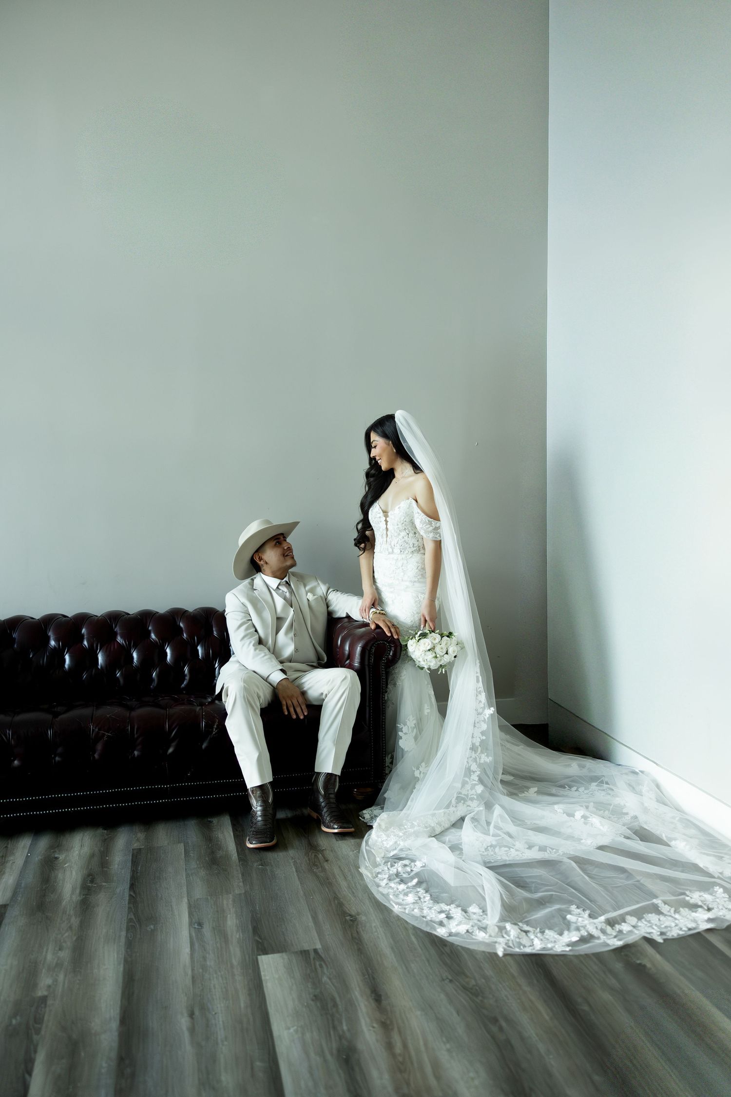 A bride in a flowing lace gown stands beside a groom in white sitting on a leather couch.
