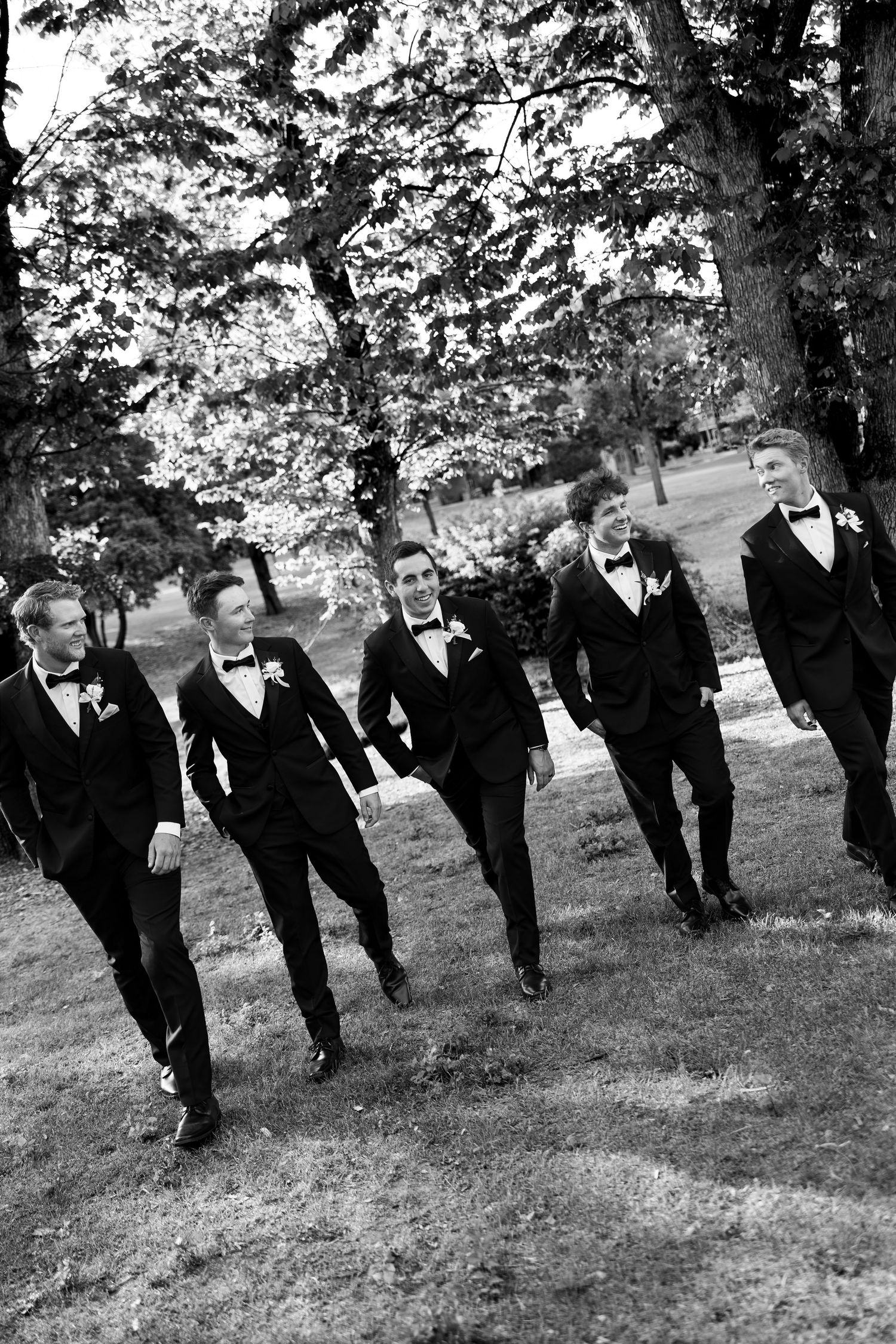 Five groomsmen in black tuxedos and bow ties walk together outdoors on grass with trees in background, black and white photo.