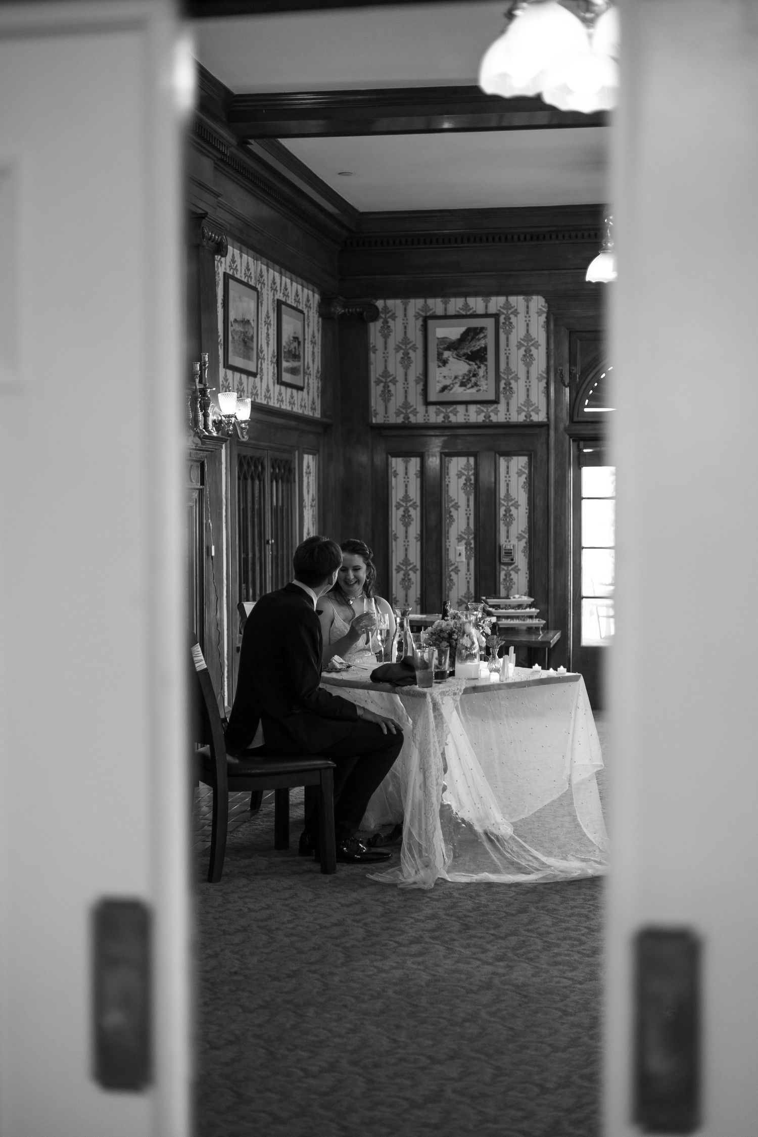 Bride and groom sit together at a sweetheart table in a classic wood-paneled reception room, in black and white.