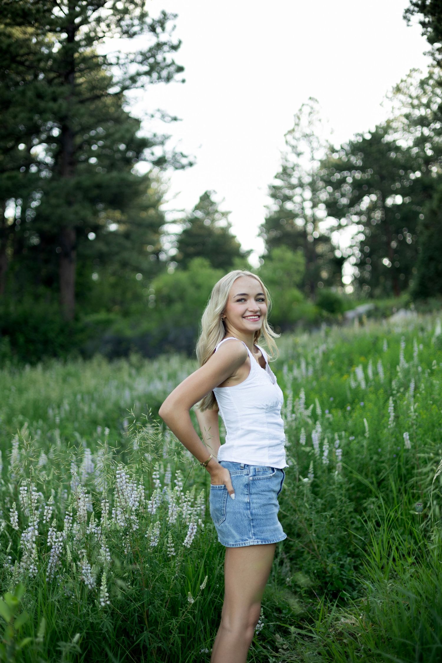 Casual summer outfit portrait among blooming lupines and tall grass with evergreen forest backdrop.