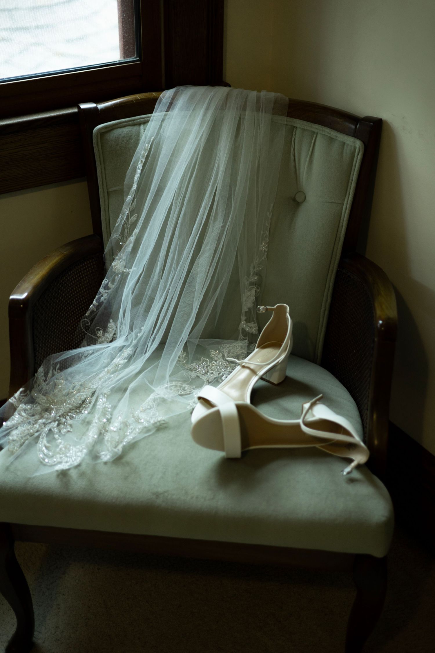 A delicate white wedding veil and bridal shoes rest elegantly on an upholstered chair near a window.