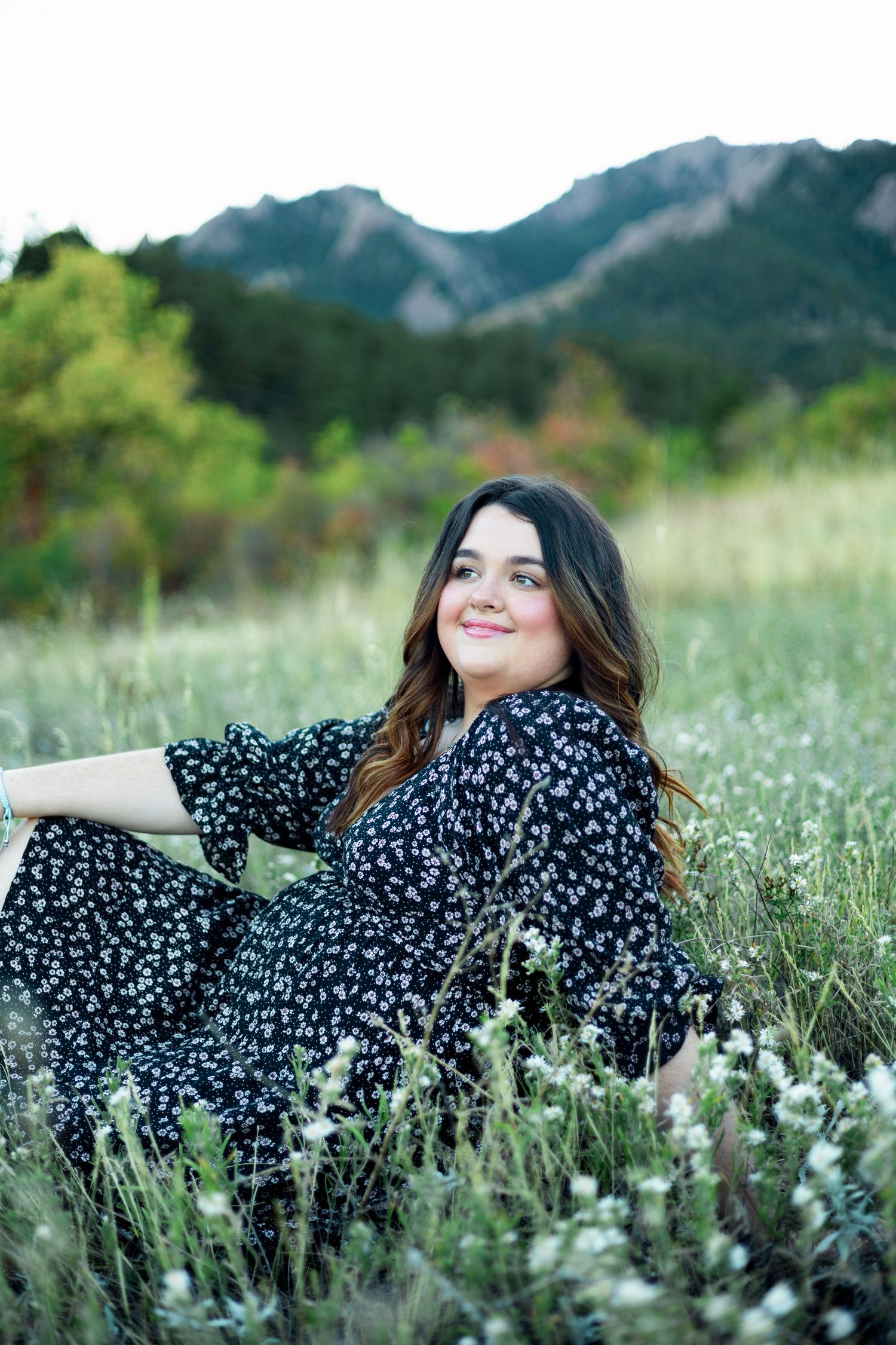 Young woman in floral dress sitting in a mountain meadow, smiling among wildflowers with rocky peaks in the background.