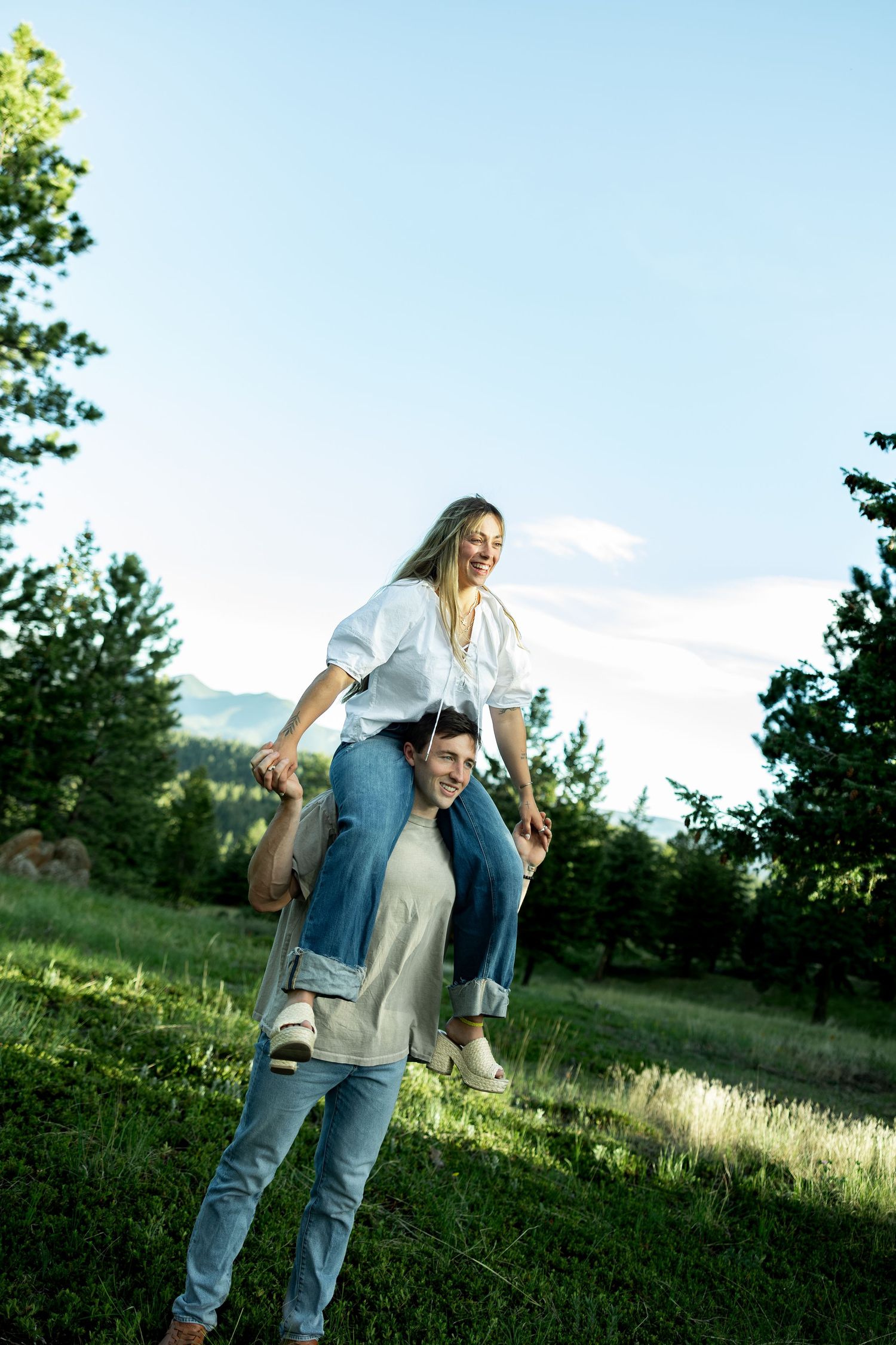 A happy couple enjoying a piggyback ride outdoors in a lush green meadow with mountains and trees in the background.