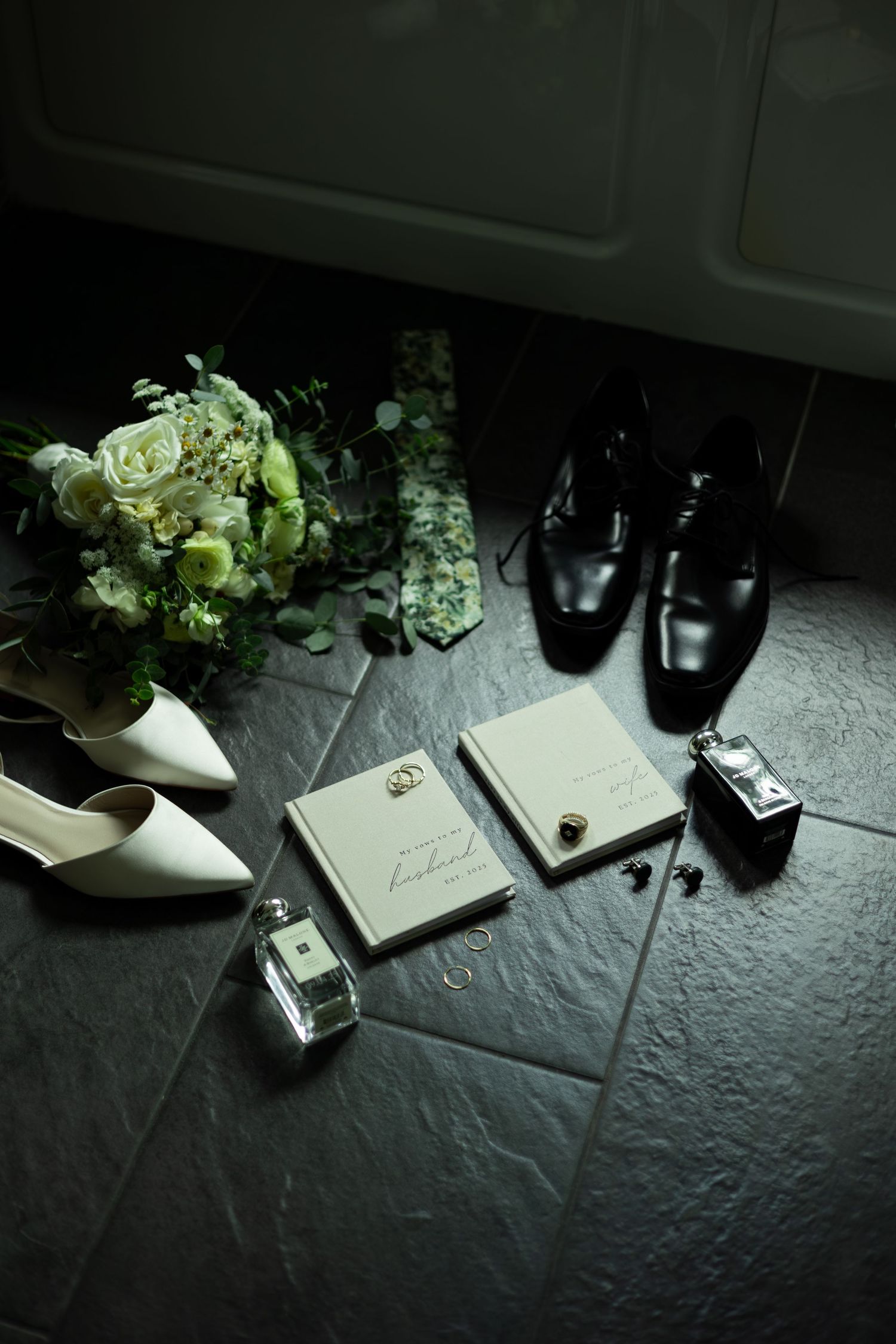 Wedding flat lay with white heels, black dress shoes, bouquet, rings, cologne, cufflinks, and vow books on dark tile floor.