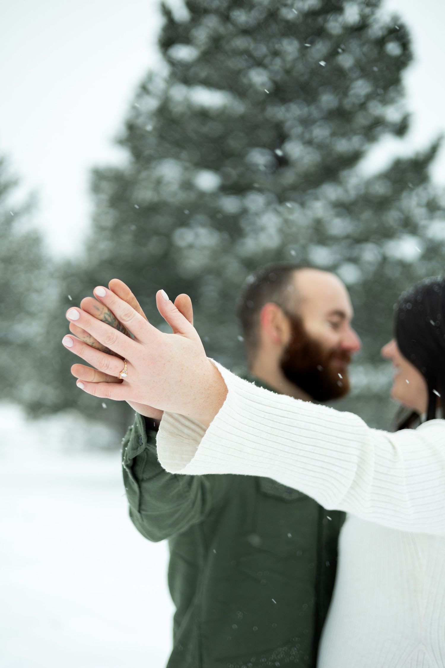 Two people share a joyful moment together in the snow with outstretched arms against a blurred evergreen backdrop.