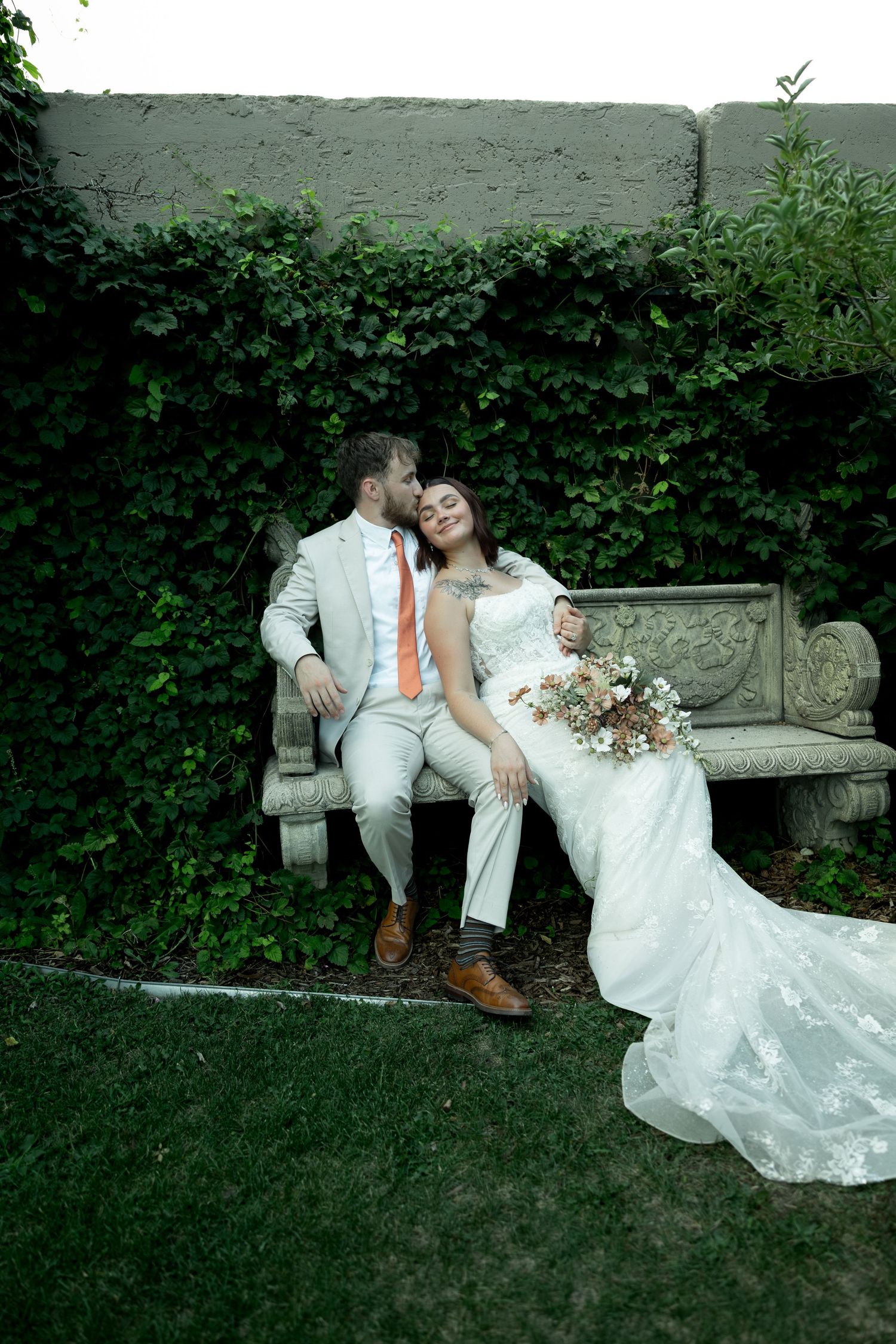 A bride and groom sit on a stone bench surrounded by lush ivy, sharing a romantic moment in wedding attire.