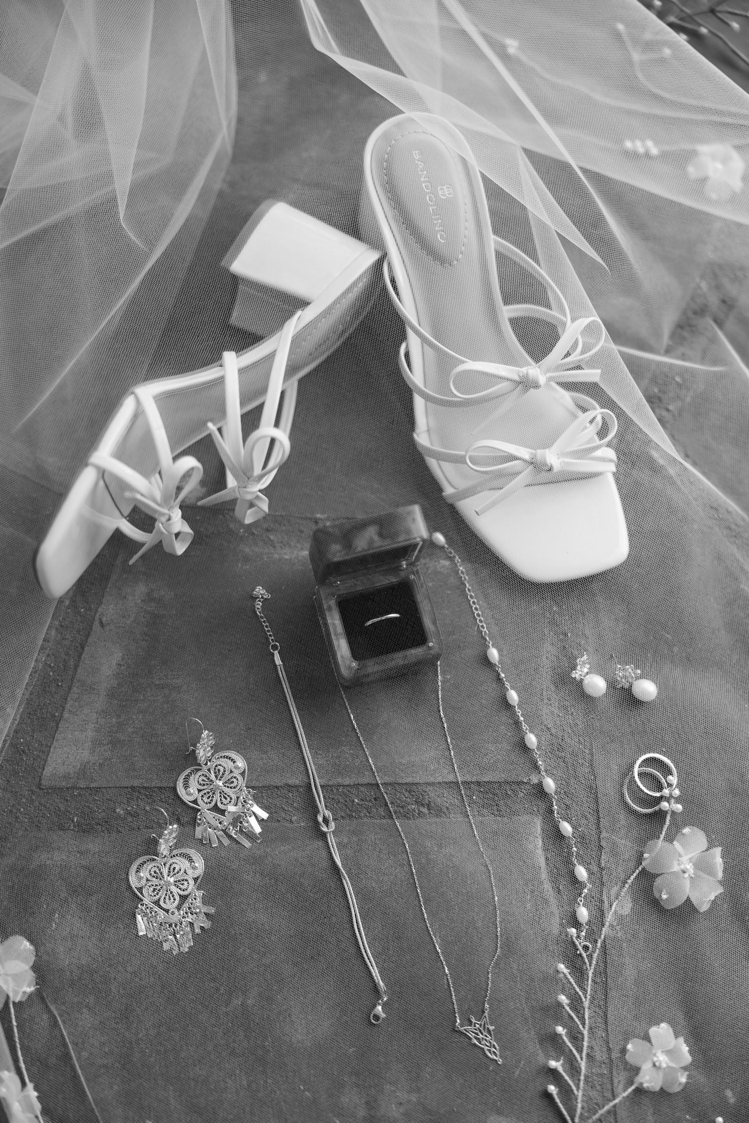 Black and white flat lay of bridal accessories including heels, ring box, necklaces, and earrings beneath a veil.