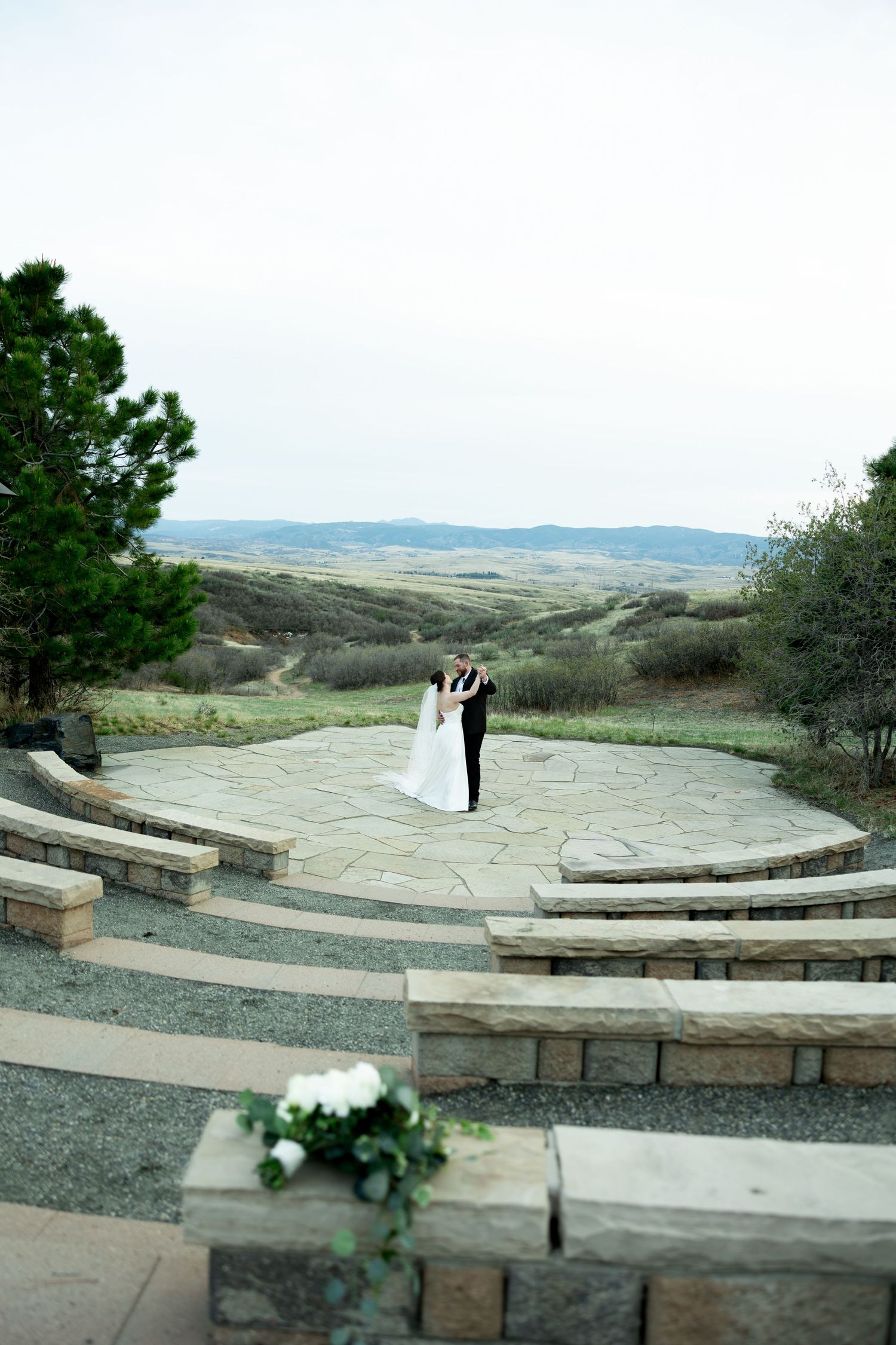 Bride and groom embrace on stone patio overlooking scenic rolling hills landscape at outdoor wedding venue.