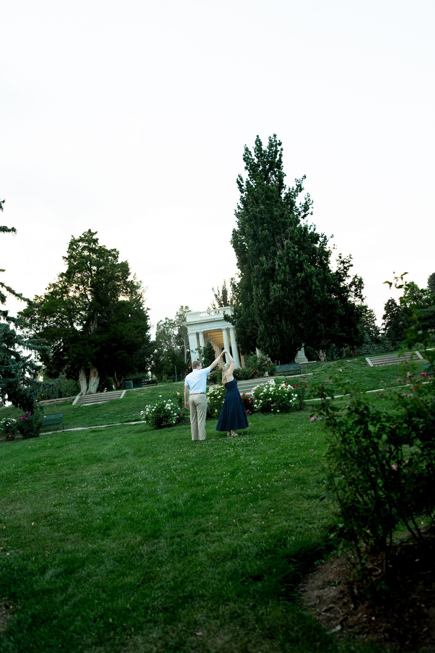 Couple holding hands on lush green lawn near white gazebo surrounded by tall trees in a serene garden park.