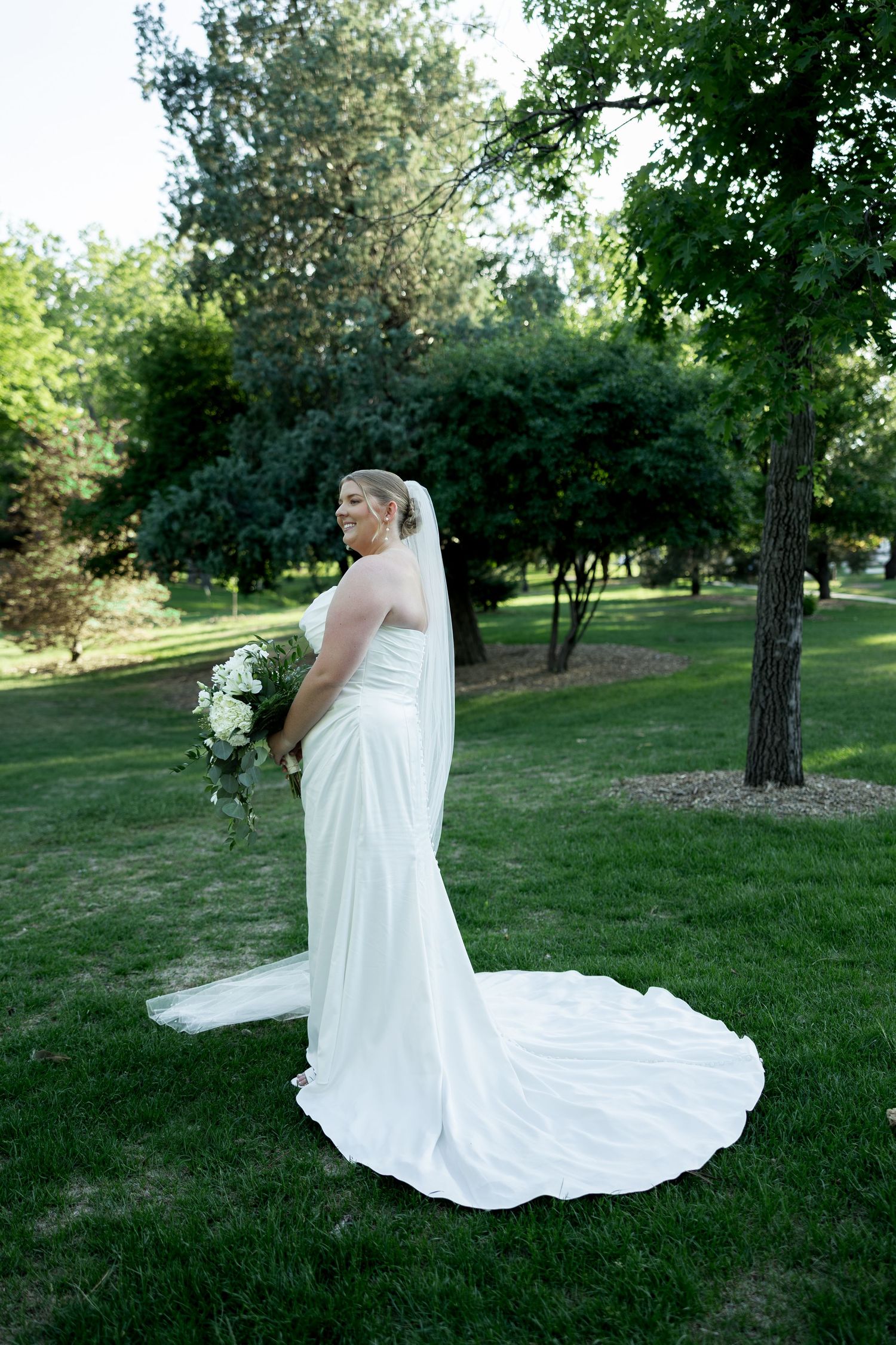 Bride poses in white strapless gown with long train and veil holding white bouquet in park.