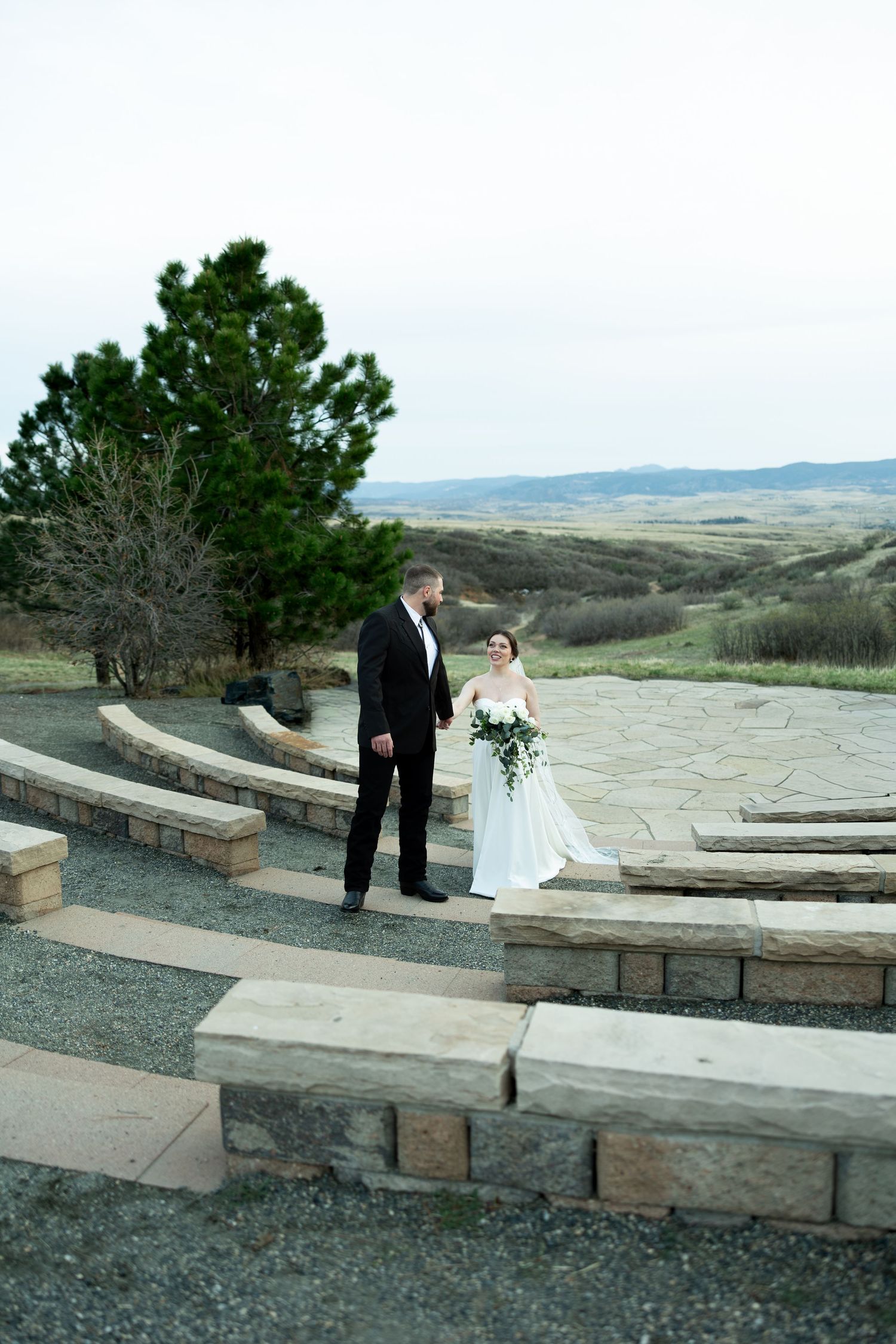 Bride and groom stand on stone amphitheater steps overlooking rolling hills, bride holding cascading white floral bouquet.