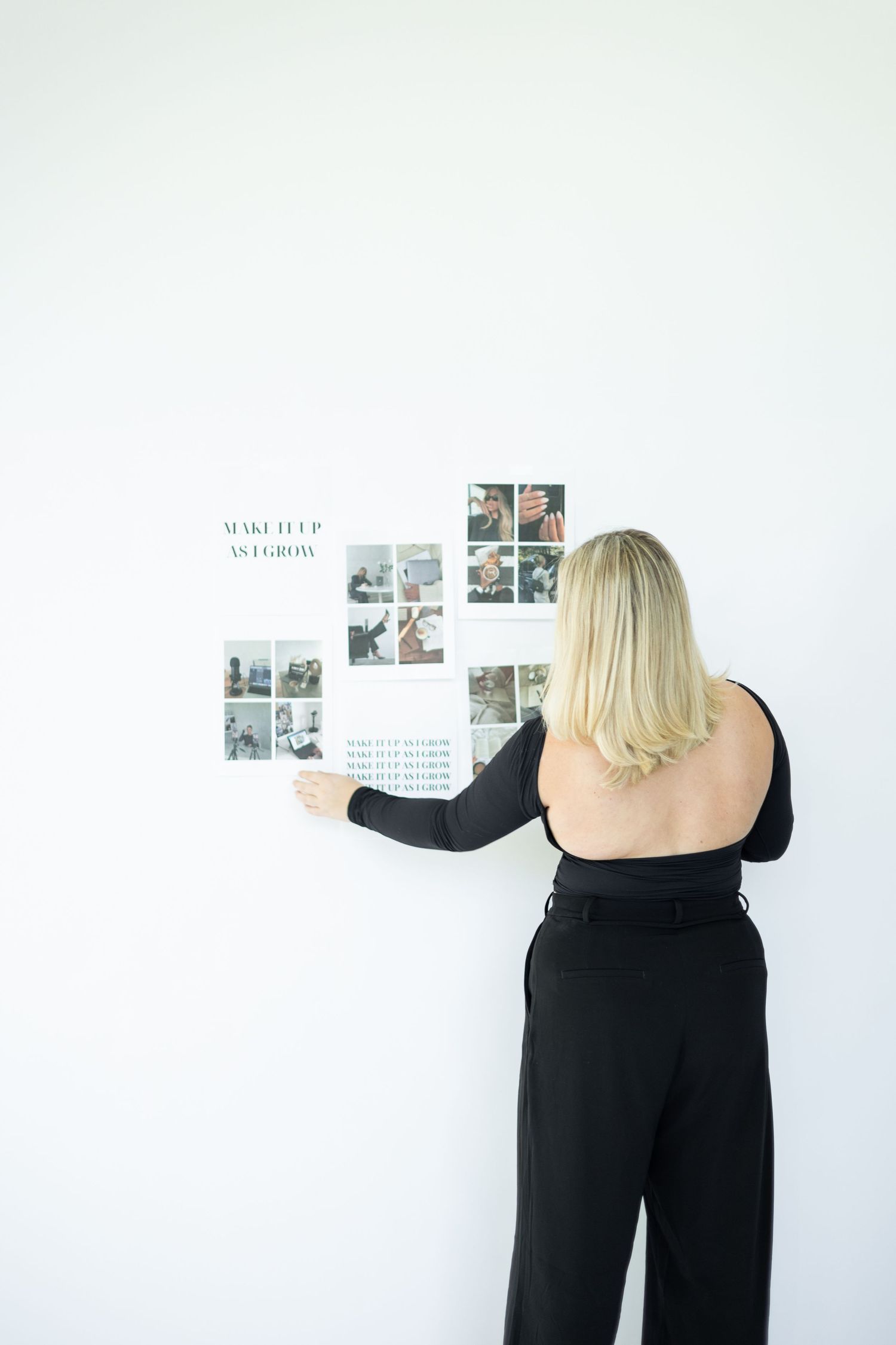 Person in black outfit arranging photos and text on white wall.