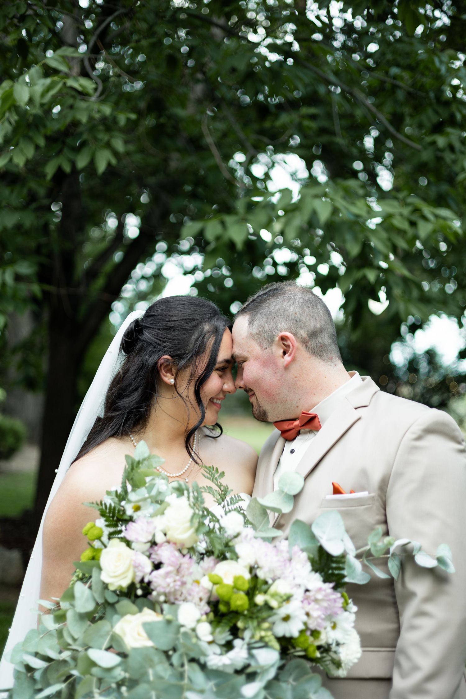 Romantic outdoor wedding portrait under green trees with a beige suit and floral bouquet.