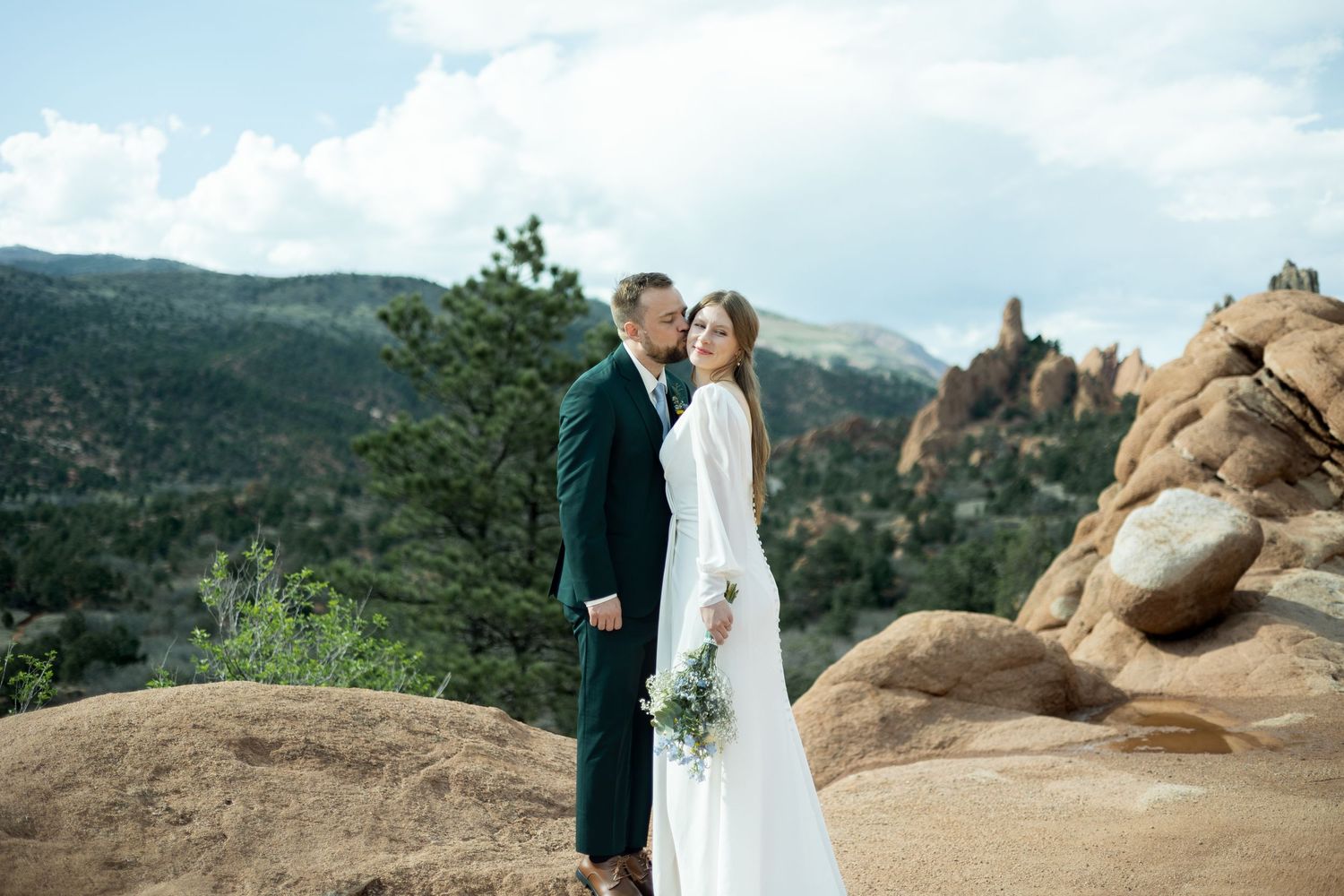 A couple in wedding attire stands together on a rocky outcrop with mountains and blue skies in the background.