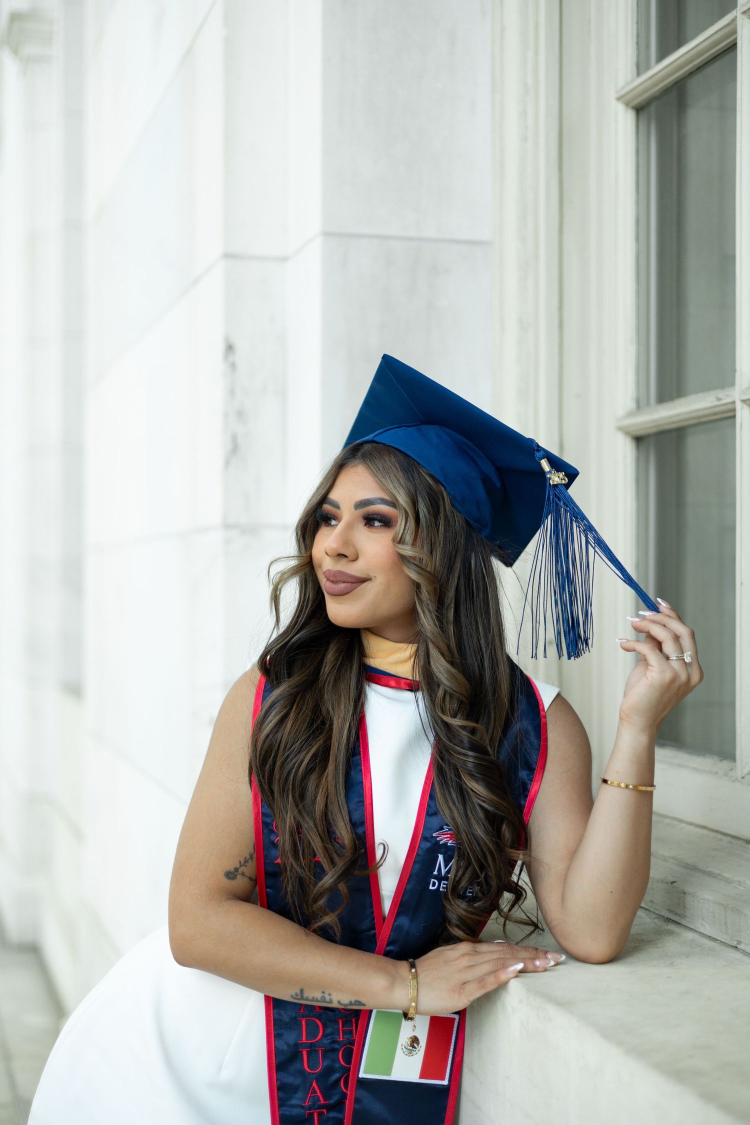 Graduate in blue cap and gown with decorative stole poses while holding tassel during outdoor graduation photo session.