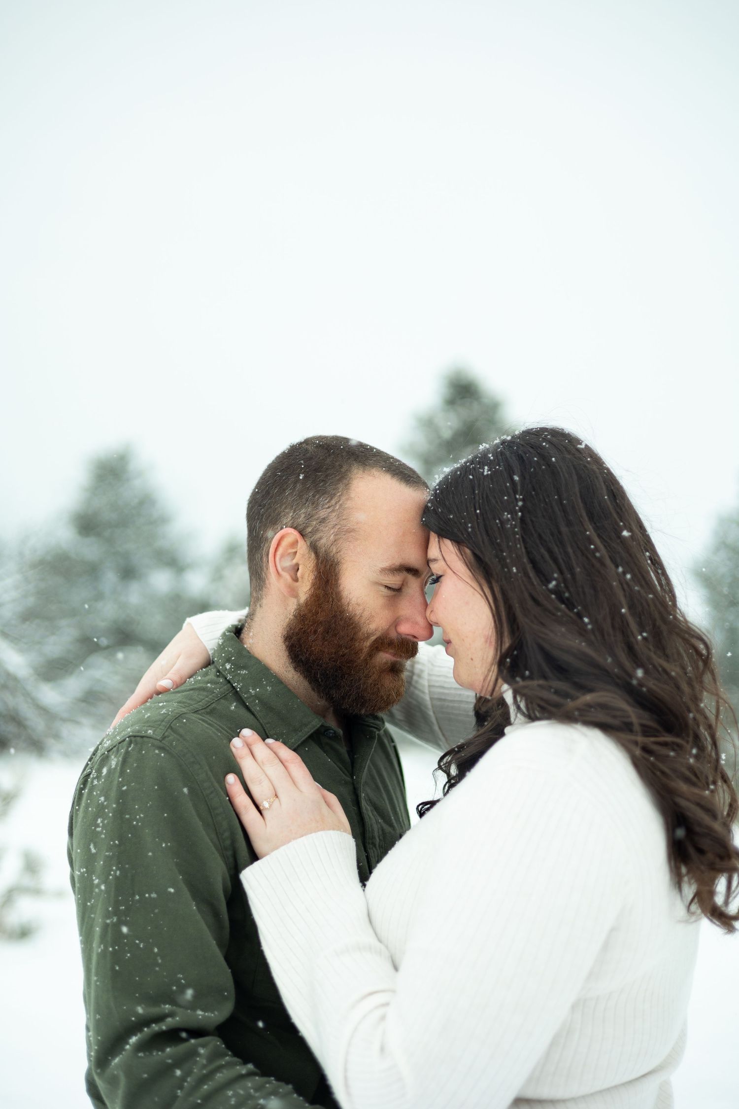 Two people in winter clothing share an intimate moment together as snow falls in a snowy outdoor setting.