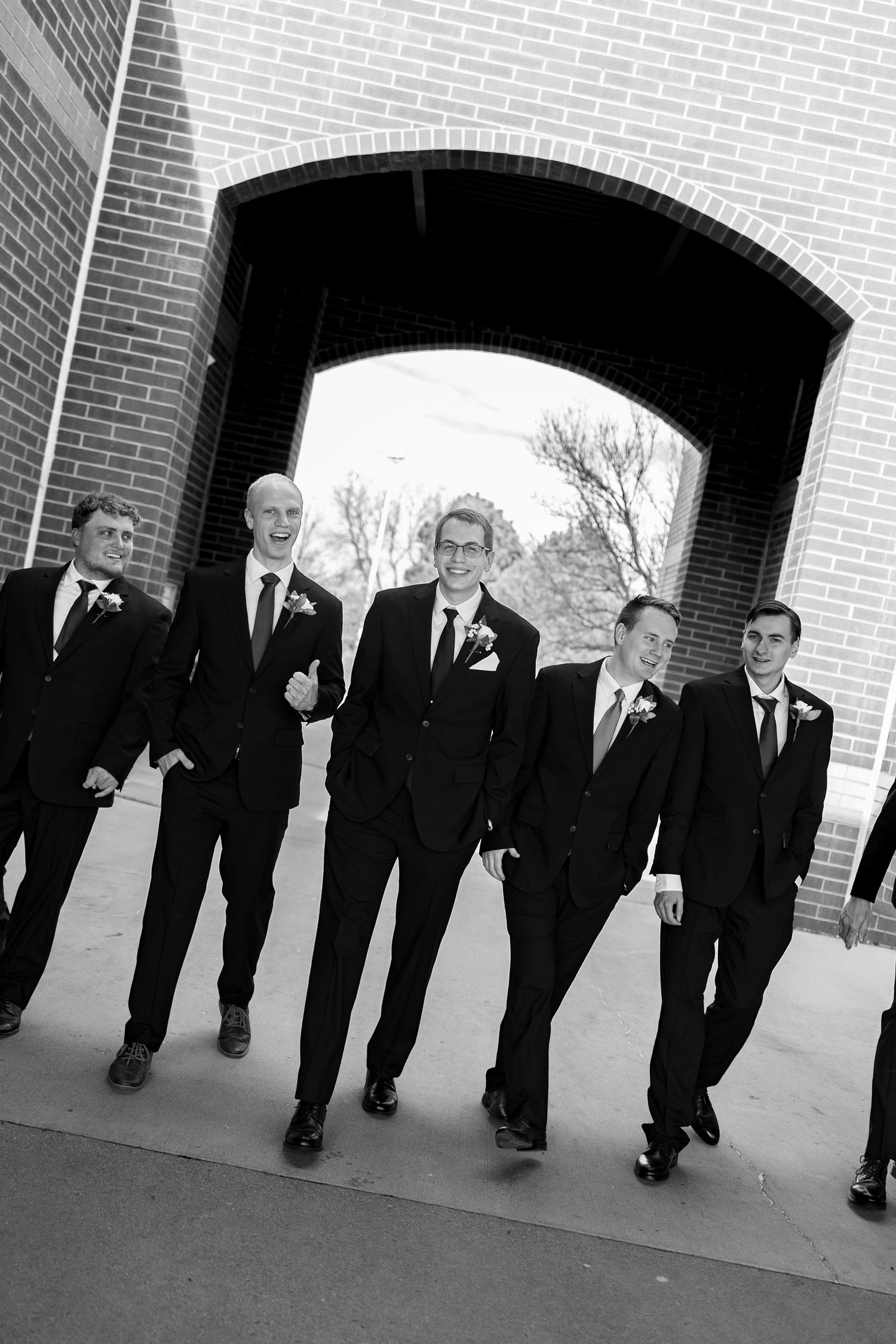 Groomsmen in black suits walking confidently under a brick archway in a classic black and white wedding photo.